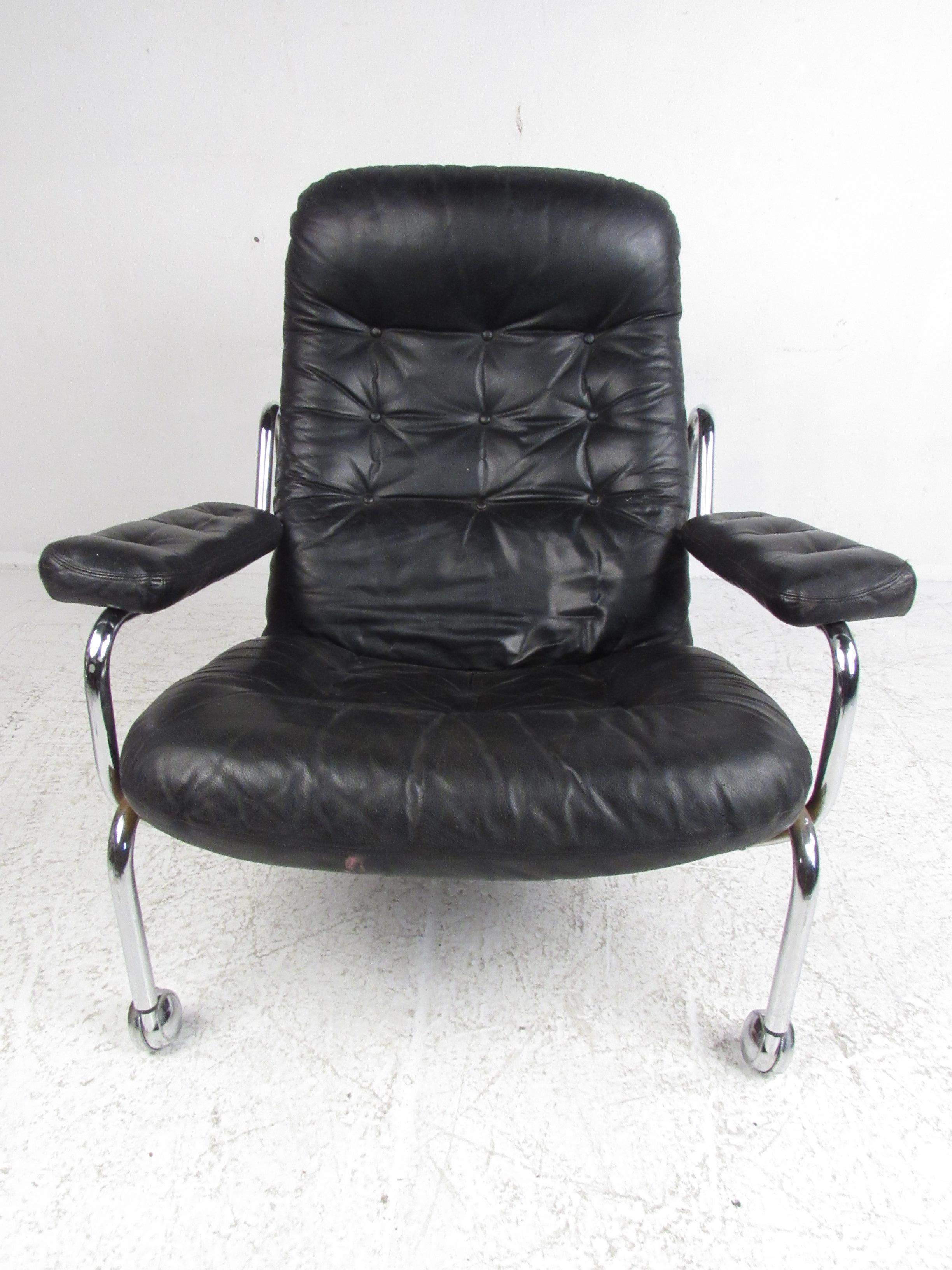 A stunning vintage modern lounge and ottoman covered in tufted black leather with a tubular chrome plated frame. This sleek lounge chair boasts thick cushioned arm rests, a sculpted frame, and castors on the front for convenience. The overstuffed