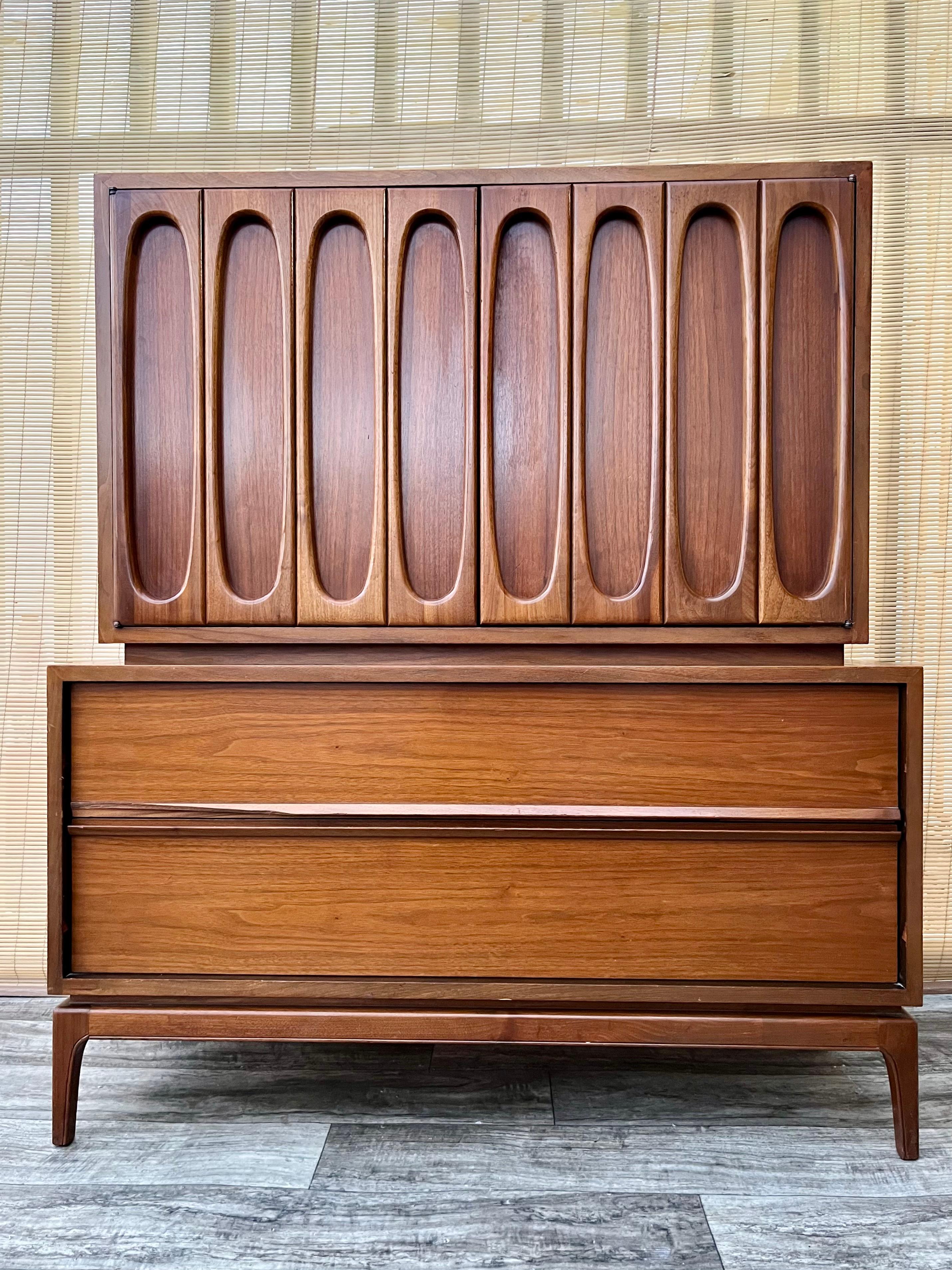 Vintage Mid Century Modern Brutalist Inspired Highboy Dresser. Circa 1960s.
Feature a monumental look with brutalist inspired designed doors, a beautiful walnut wood grain finish, and five drawers offering plenty of storage space. 
In good original