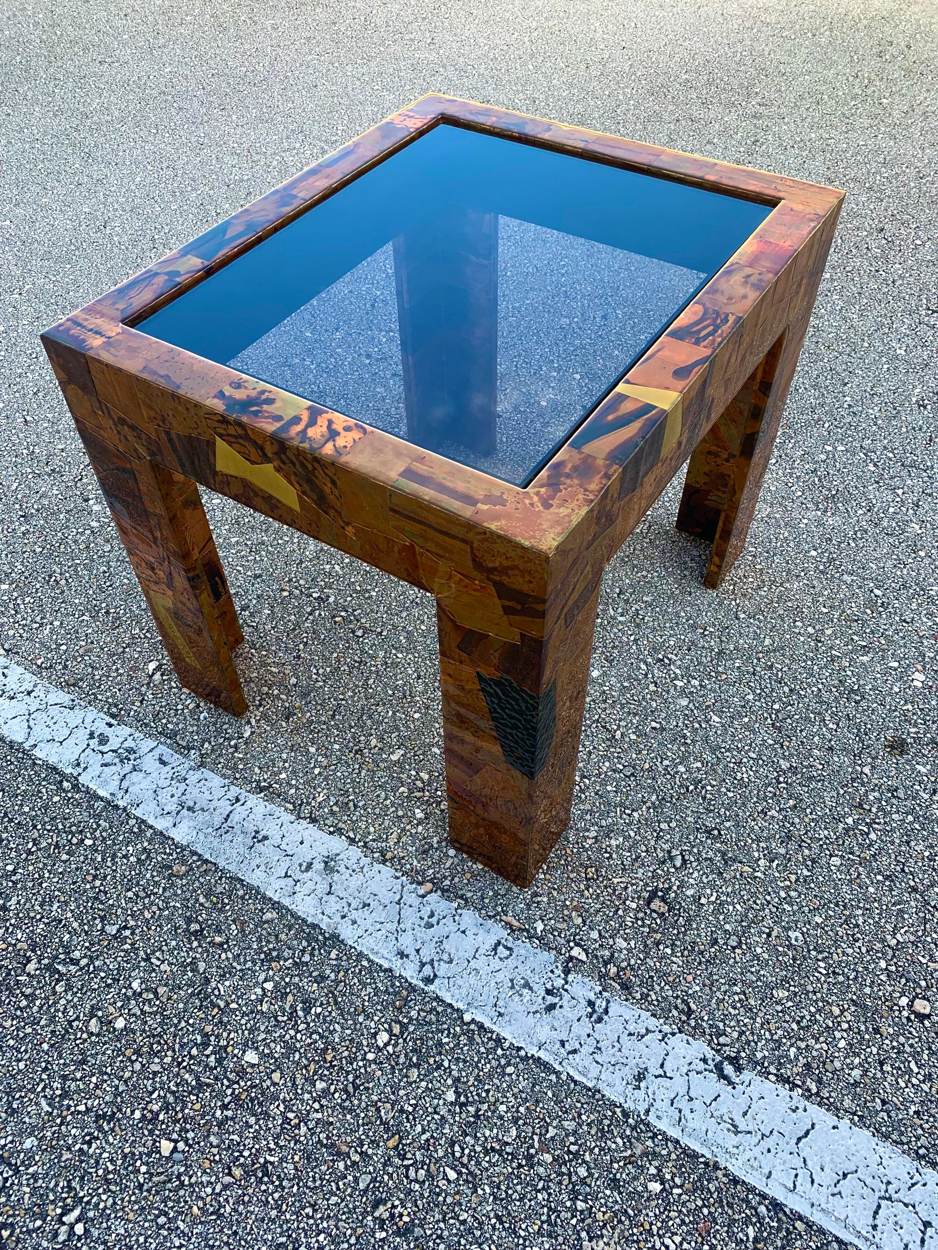 Studio made copper and brass accent table with smoked glass. Made to have a brutalist flair, the artist used different pieces of textured copper and brass to cover the entire table. The plates are affixed with copper staples then coated in a thick