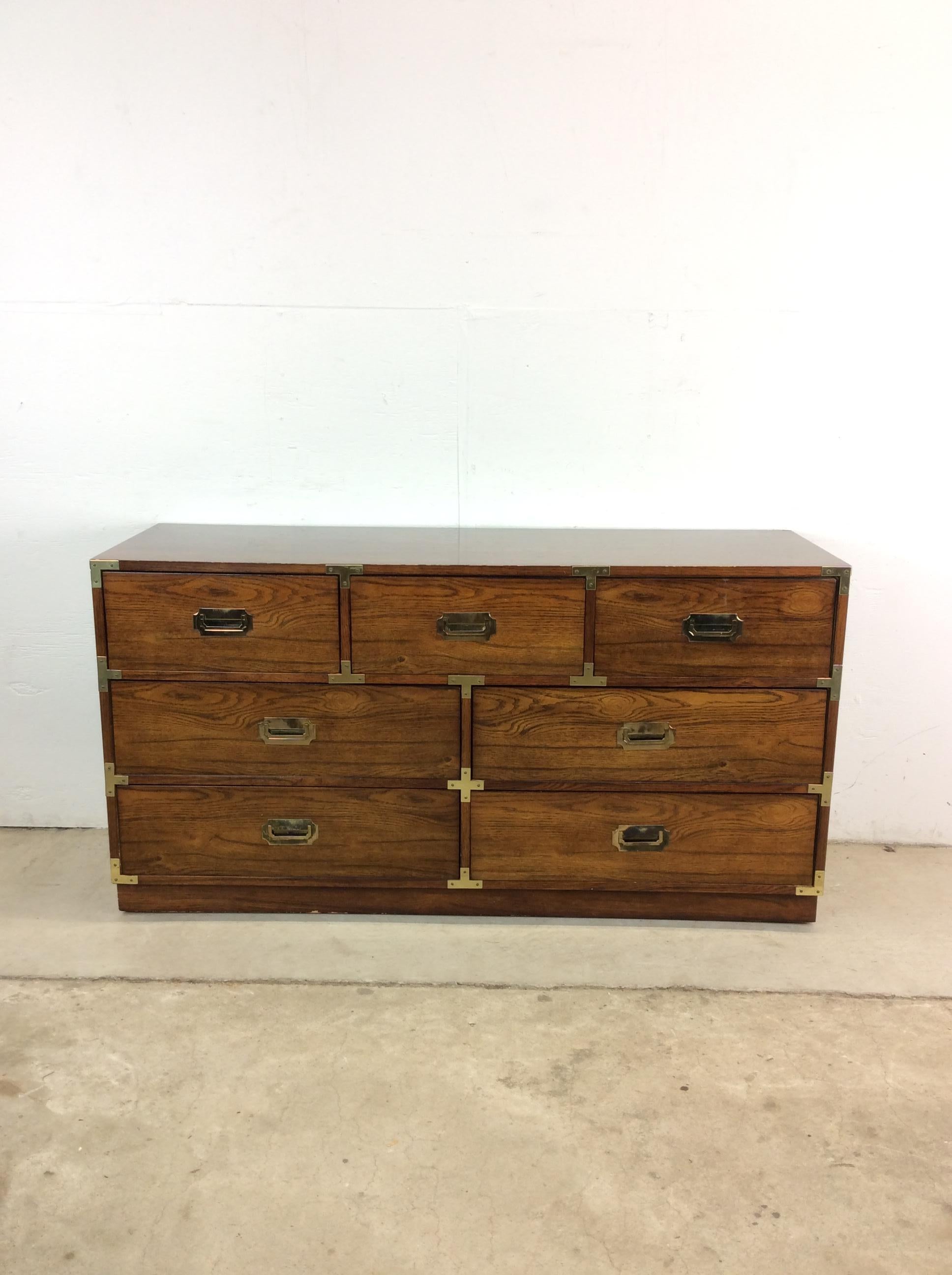 This mid century campaign style dresser features hardwood construction, oak veneer with original finish, and seven dovetailed drawers with brass accented hardware.

Dimensions: 56.25w 19.25d 29.5h

Condition notes: Original finish is in good