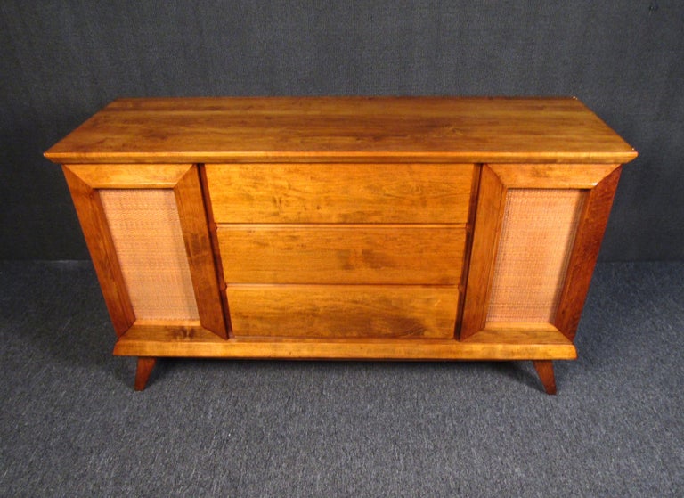 A vintage server full of Mid-Century Modern style, this piece can a brighten a room with its warm woodgrain and cane accents. With two side compartments and three large drawers, this server offers plenty of space for storage. Please confirm item