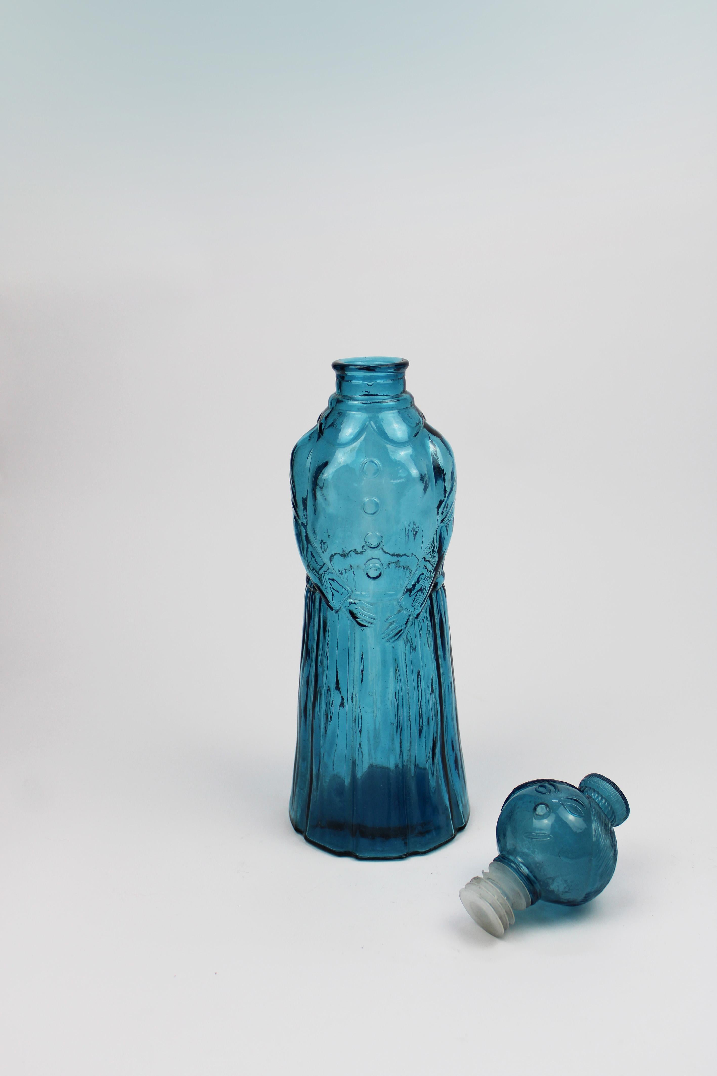 Questa sorprendente caraffa moderna di metà secolo, risalente agli anni '60, è realizzata in vetro blu goffrato a Empoli, in Italia. Sormontato da un tappo a forma di donna sorridente, il pezzo emana una giocosa raffinatezza. Esemplificando il vetro