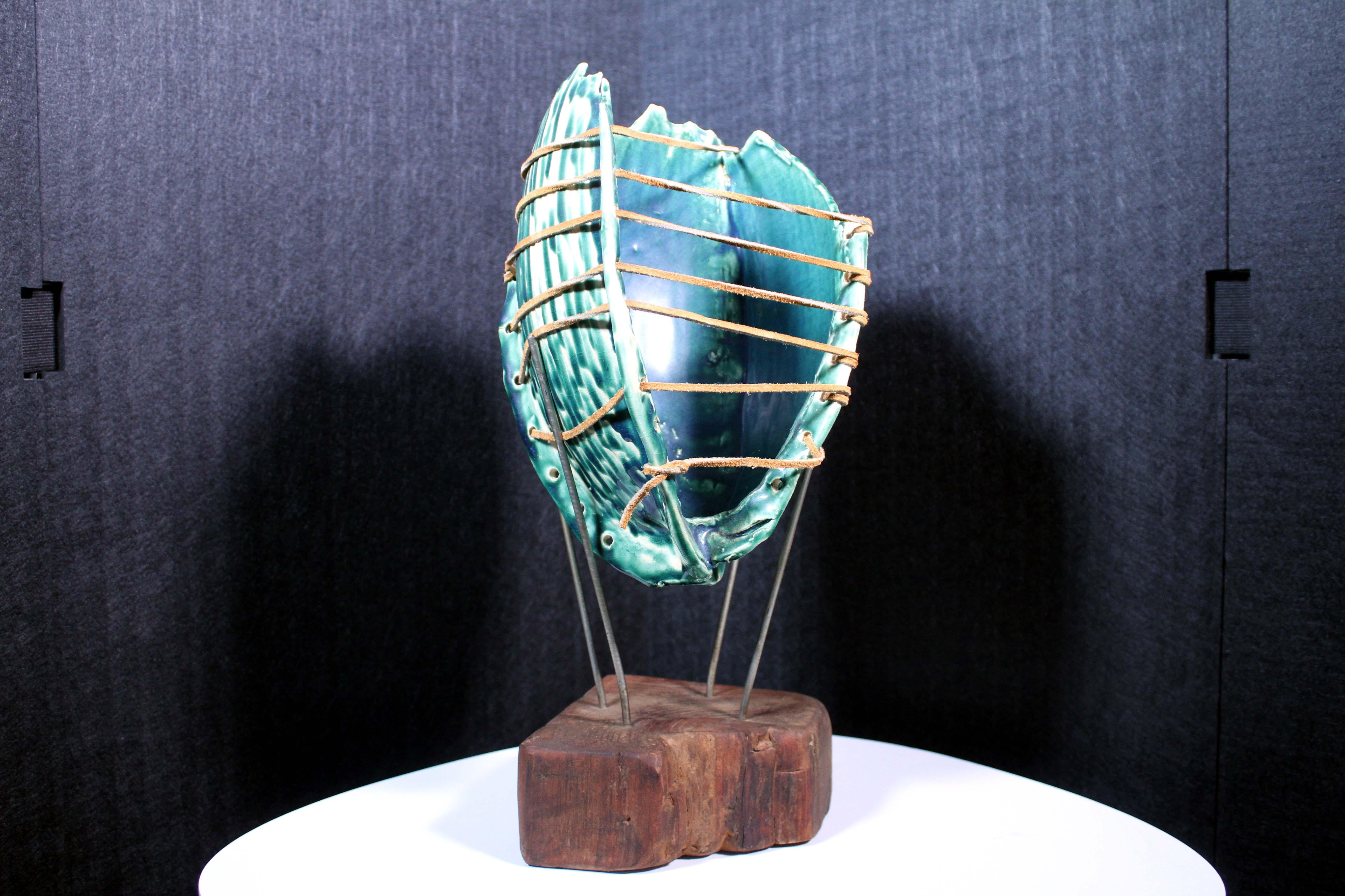 A unique Mid-Century Modern decorative ceramic item showcasing a biomorphic shell design. The sculpture has lovely tones of swirling green with leather strands. It sits upon a metal stand attached to an organic wood base. A marvelous statement and