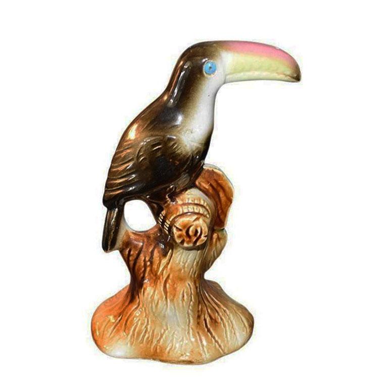 A painted ceramic bird figurine. Made in Brazil, this small statue is of a toucan, in black, sitting upon a brown tree branch. His beak is in yellow and pink, and his eyes are painted blue. A fabulous MCM piece perfect for decorating any bookshelf