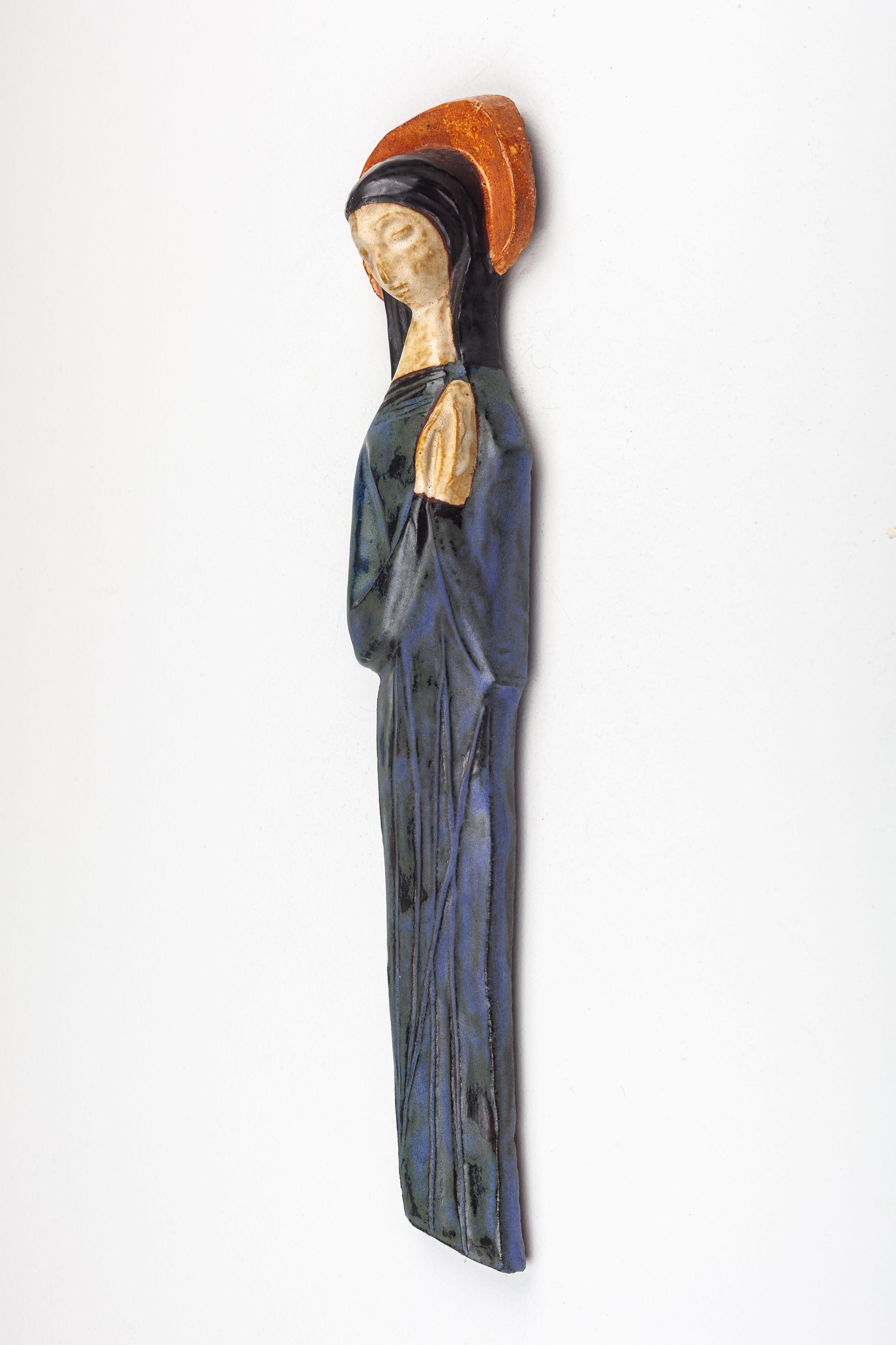 This mid-century modern ceramic sculpture portrays the Virgin Mary, a central figure in Christian iconography, handcrafted with a unique artistic interpretation by a European studio pottery artist. The figure is rendered with stylized simplicity,