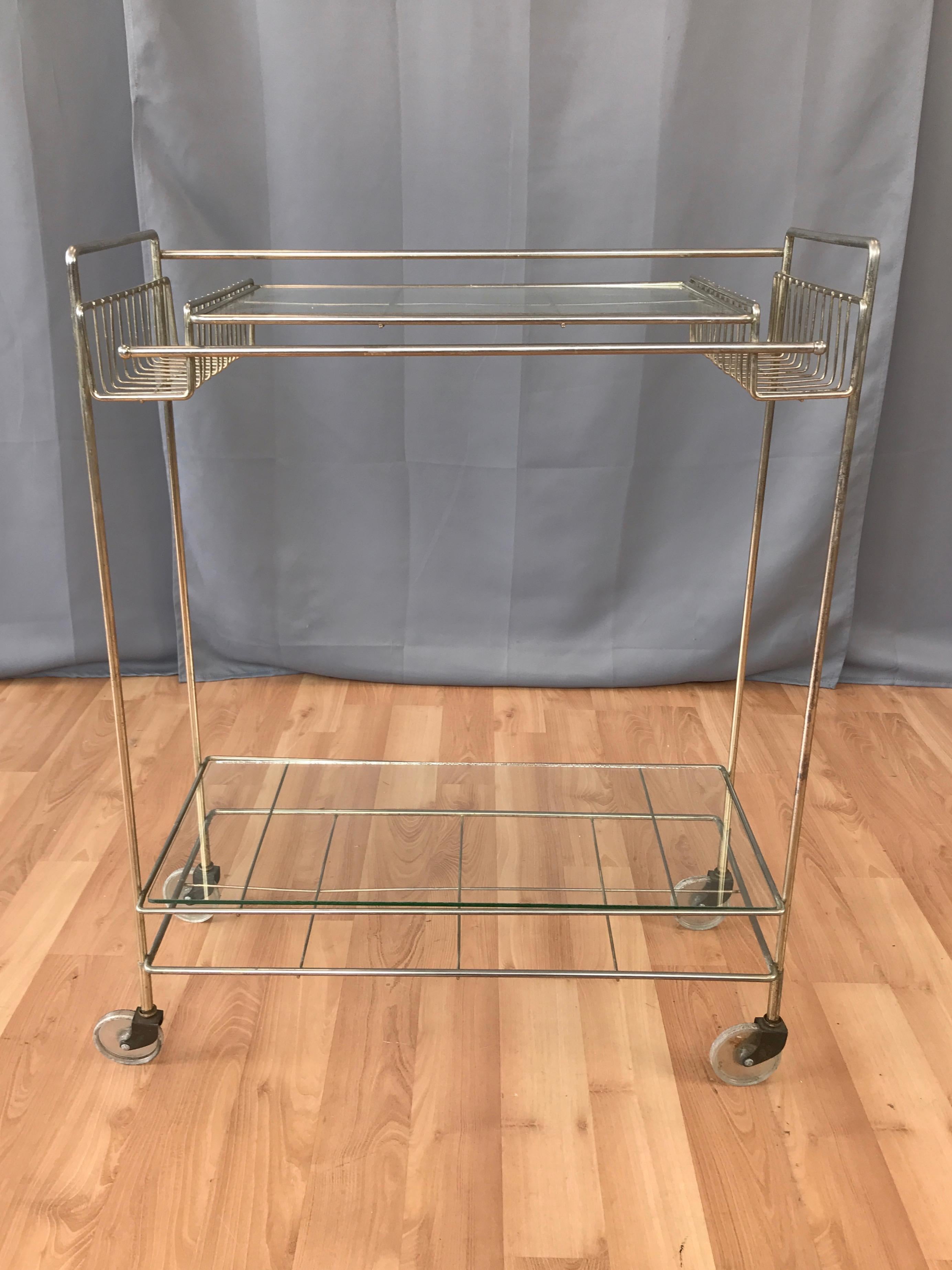 A visually light Mid-Century Modern two-tier metal and glass bar or serving cart from the 1960s.

Minimalist, barely there frame of slender steel rods with a pale champagne finish. (Appears a bit less brassy in person; image 11 is closest.) Baskets