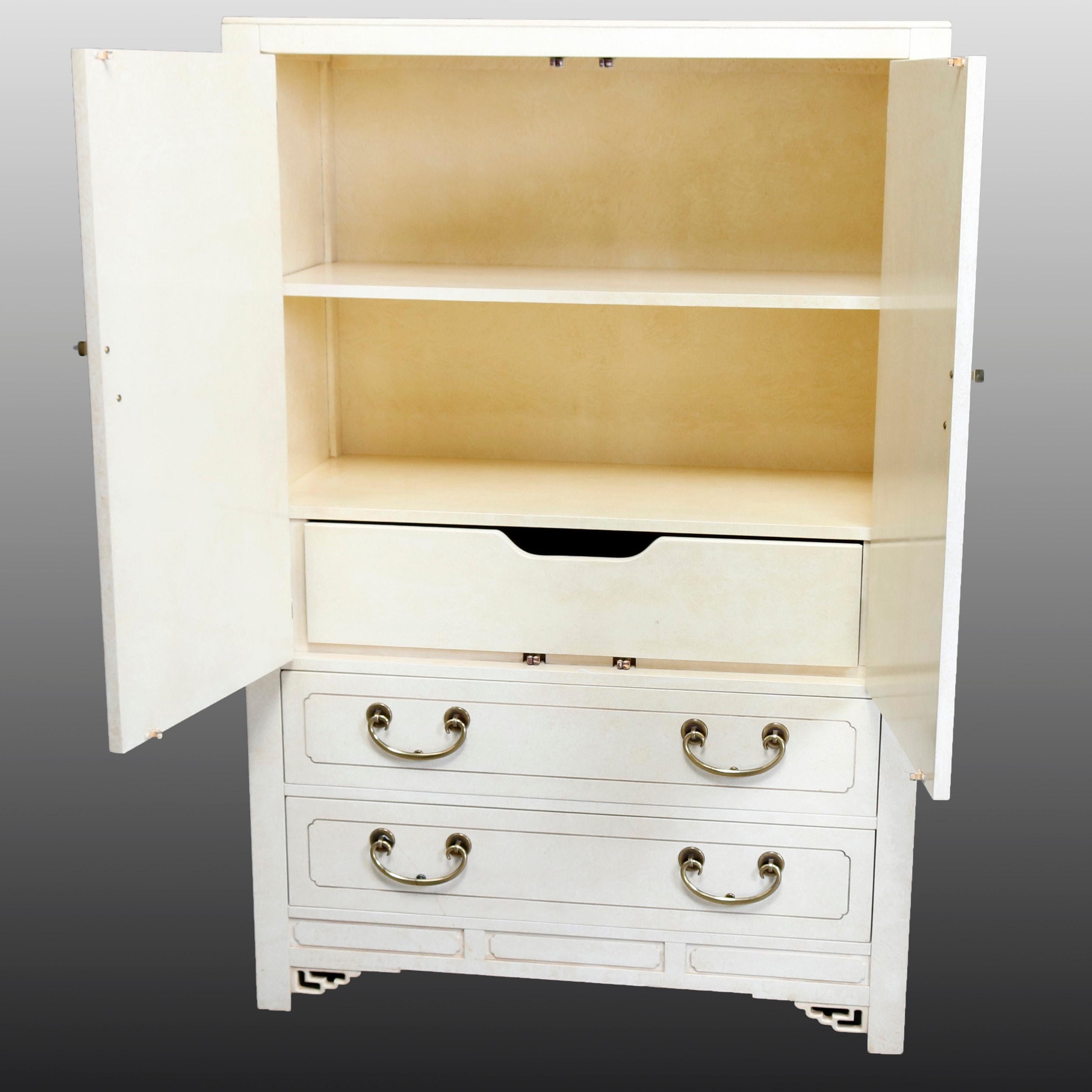 A Mid-Century Modern wardrobe or linen press by Hickory White offers Chinese Chippendale styling with all-over ivory enameled finish, pierced corbels having Asian influence, and cast brass hardware and accoutrements. Case with upper double doors