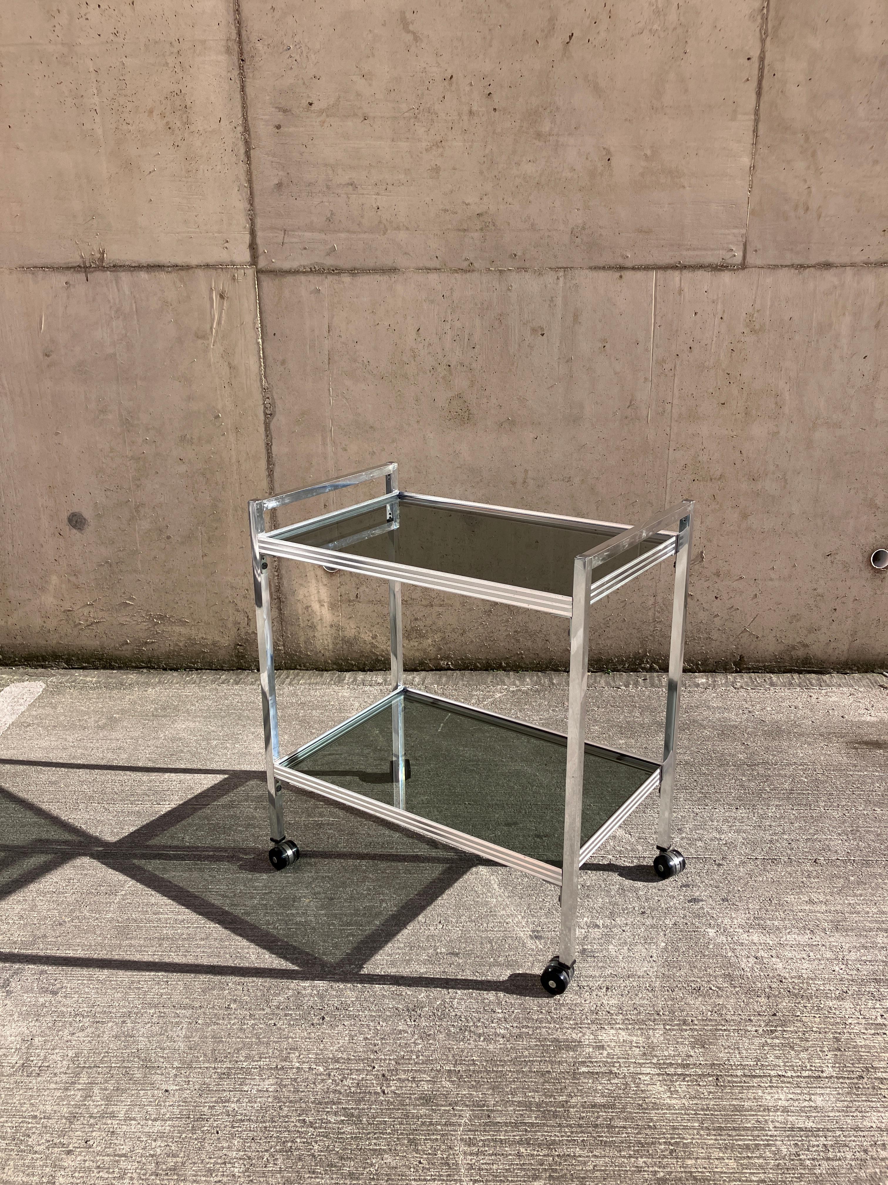 Un superbe chariot à boissons à deux niveaux du milieu du siècle dernier en verre gris et en chrome. Un design angulaire saisissant. 

Très bon état pour l'âge avec quelques petites marques liées à l'âge.

Dimensions totales : 65 cm x 41 cm x 73 cm