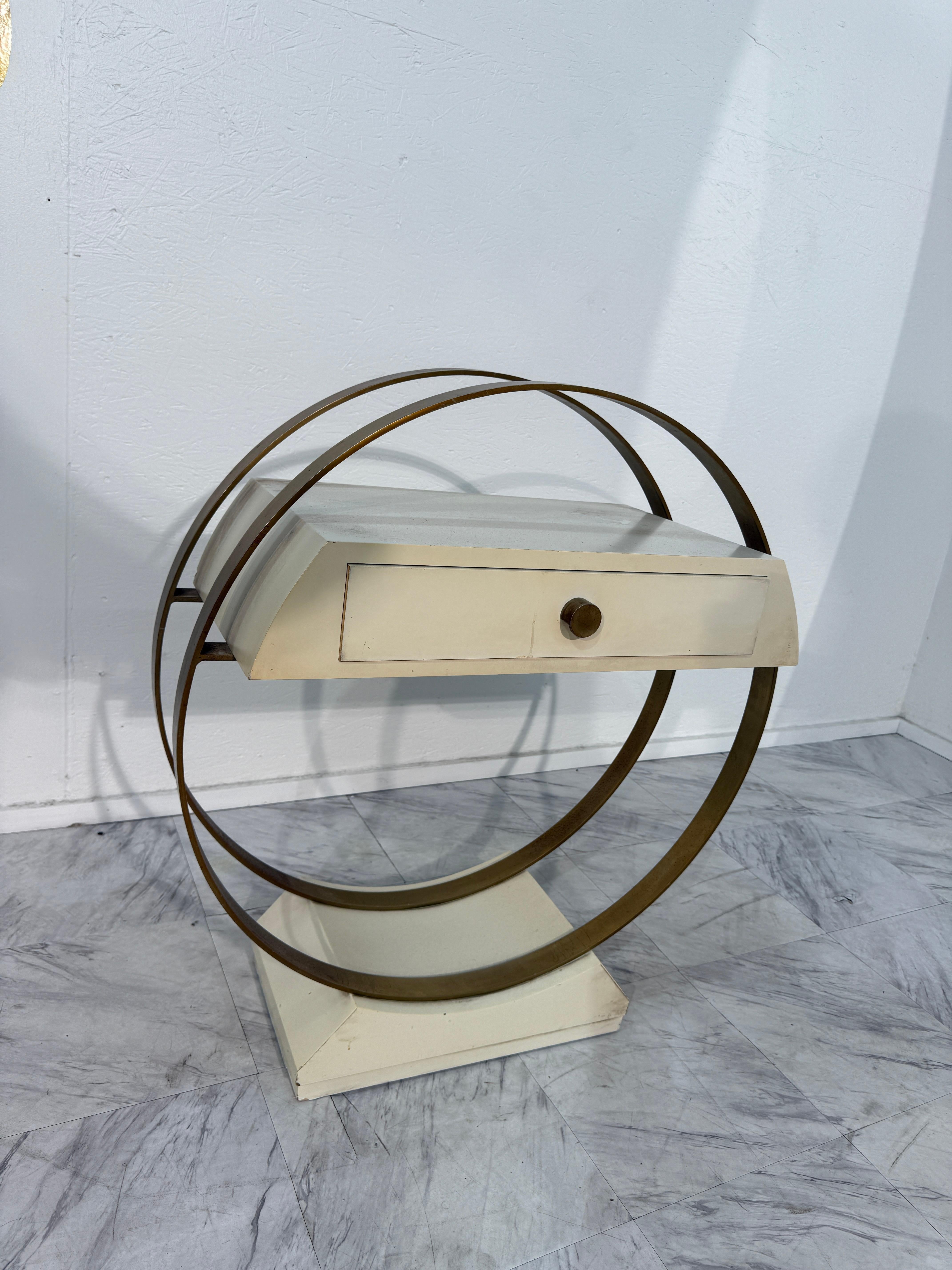 Tavolino scultoreo e futuristico nello stile del design italiano del Mid-Century Modern, caratterizzato da una sorprendente struttura circolare in ottone che abbraccia una cassettiera laccata color crema. Il gioco geometrico di curve e linee rette