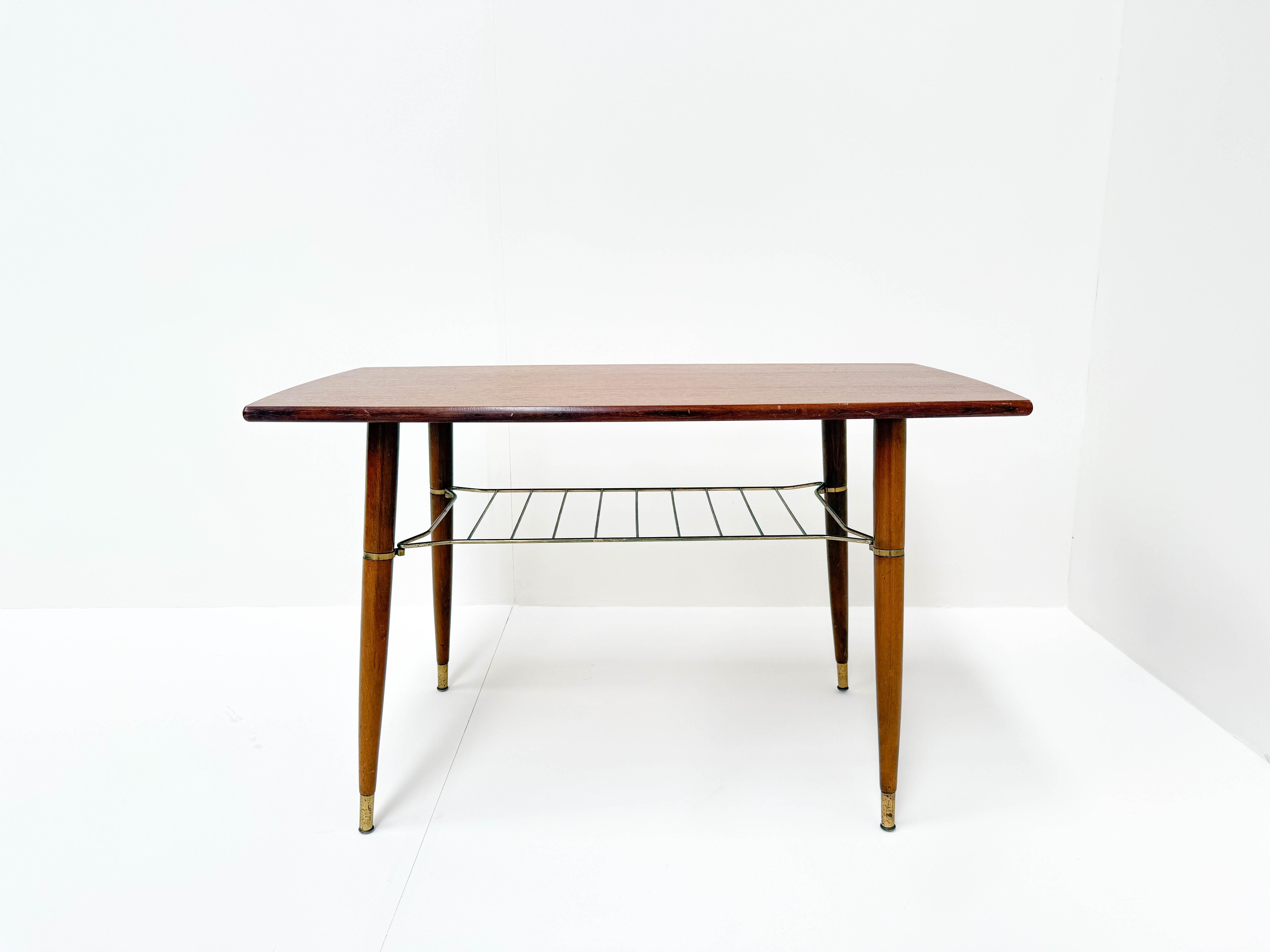 This elegant mid-century modern coffee table features a refined teak wood top with softly curved edges, embodying a timeless Scandinavian aesthetic. The table stands on four slender, tapered wooden legs with brass-capped feet. A sleek, gold-toned