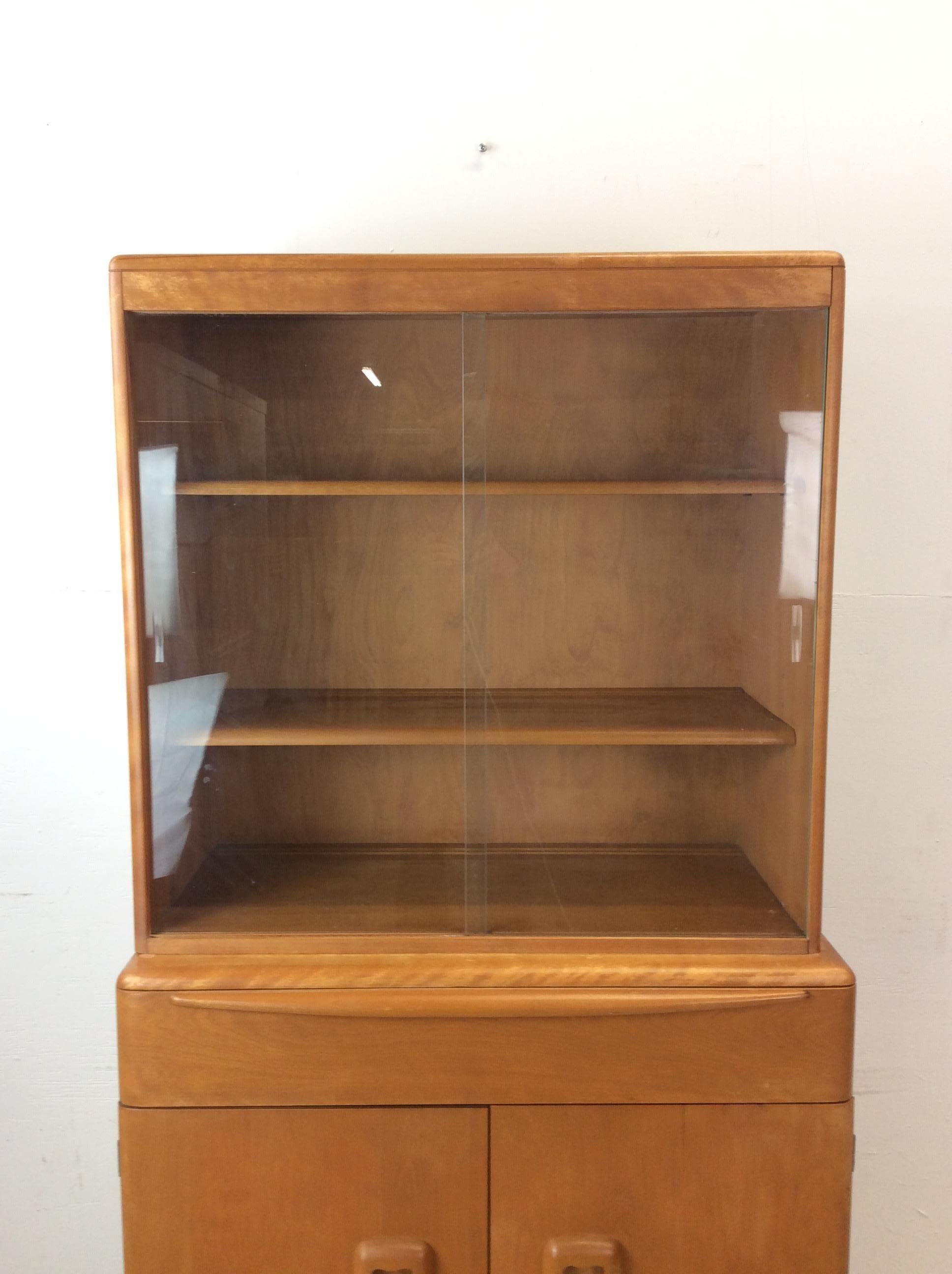 This mid century modern compact china cabinet by Heywood Wakefield features hardwood construction, original champagne finish, two piece design, glass sliding doors and adjustable shelf up top, single drawer and two door cabinet with adjustable shelf