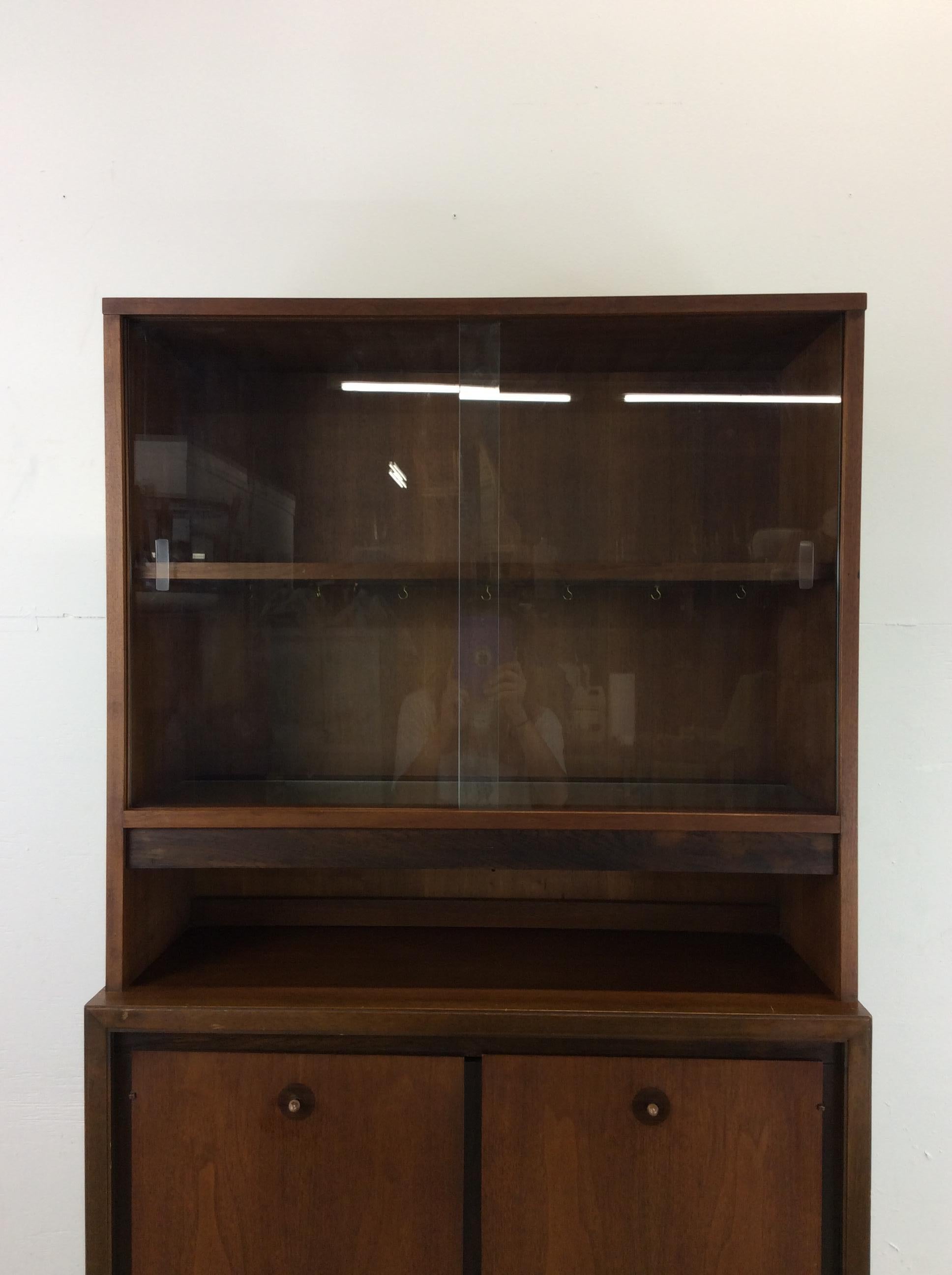 This mid century modern compact china cabinet by Basic-Witz features hardwood construction, original walnut finish, two cabinet doors with brass accented hardware containing a single dovetailed drawer and open storage, sliding glass doors up top