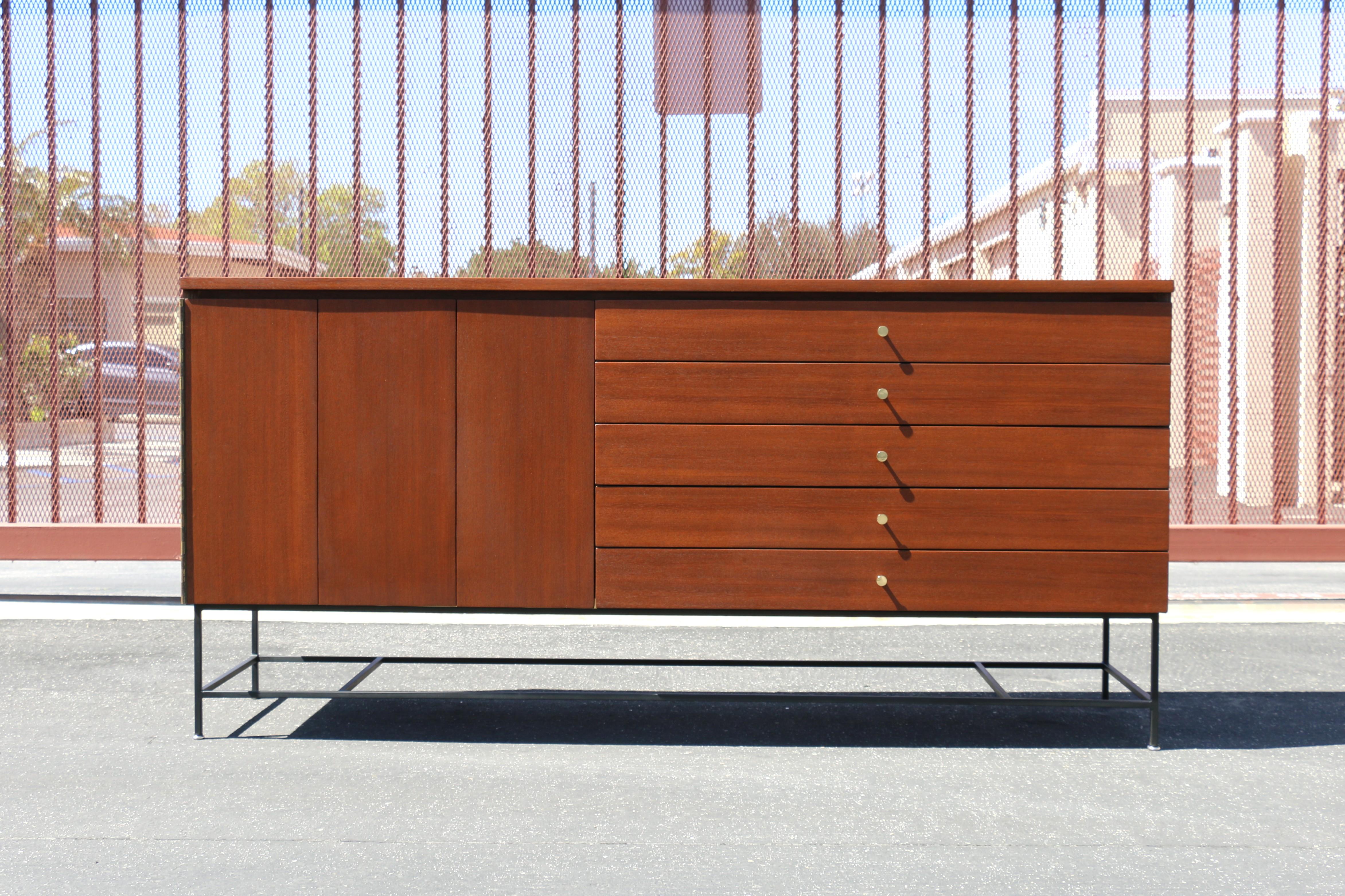Iconic Mid-Century Credenza designed by Paul McCobb for his Irwin Collection, it has the label in the first drawer. This great piece is made of mahogany wood grain with accenting brass hardware and metal frame base. Left side has a folding door and