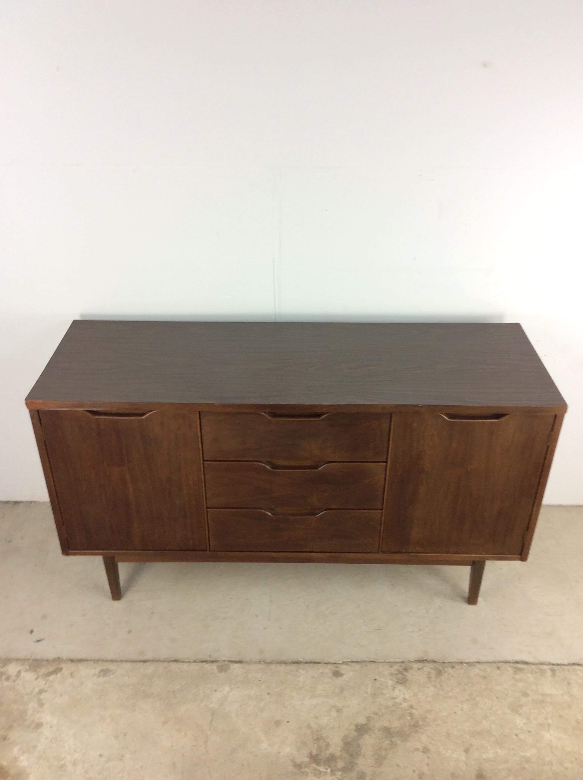 This mid century modern credenza by Stanley Furniture features hardwood construction, walnut veneer with original finish, durable formica top with faux woodgrain, three dovetailed drawers with carved wood pulls, two cabinets with single shelves and