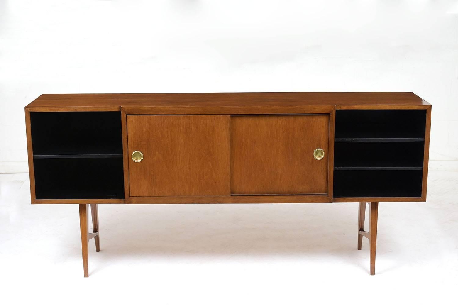 This 1960s Mid-Century Modern credenza is stained in a rich walnut color with a polished finish. The two open sections on the ends are finished in black and have shelves for storage. The centre section has two sliding doors with brass hardware and a