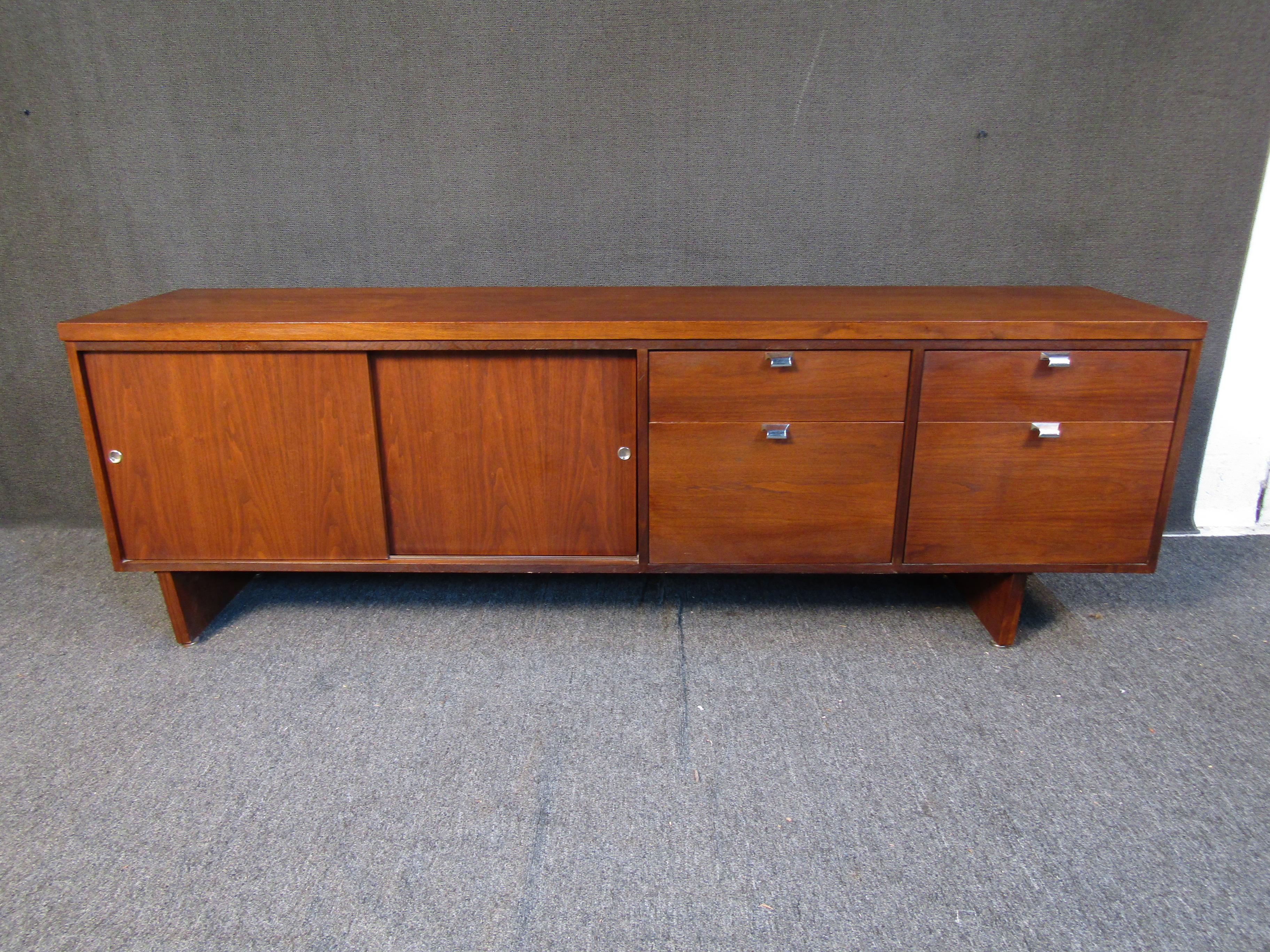 A Mid-Century Modern walnut finish credenza featuring adequate storage space and long one piece legs. The credenza also features a sliding door covering a two-tier shelving unit and 4 drawers to the right. Please confirm item location with seller
