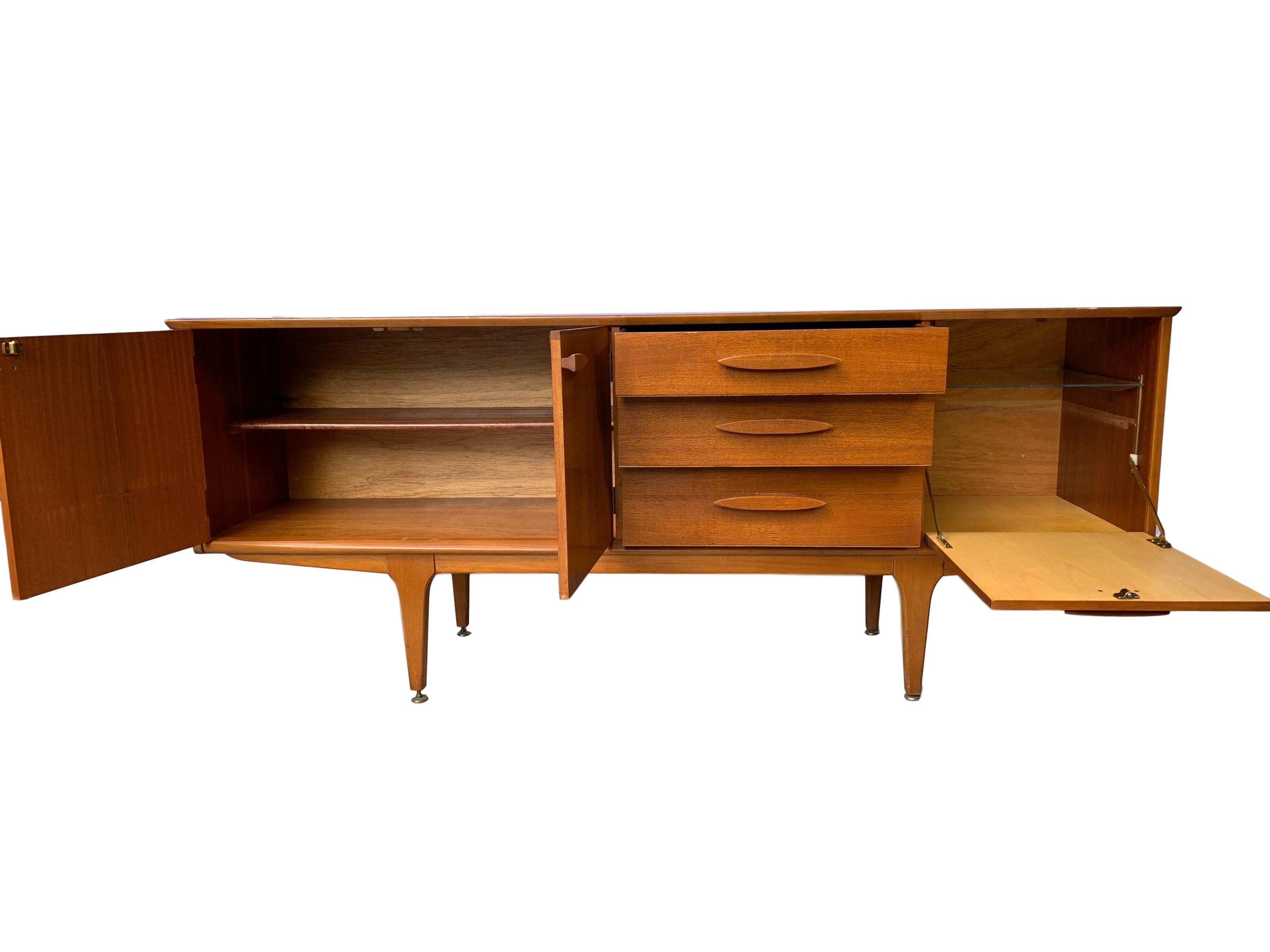 Mid-Century Modern credenza sideboard, English, circa 1960. In solid teak and flamed teak with fitted interior for loads of storage, including felt lined silverware drawer, with adjustable solid brass levelers to feet. Built to the highest