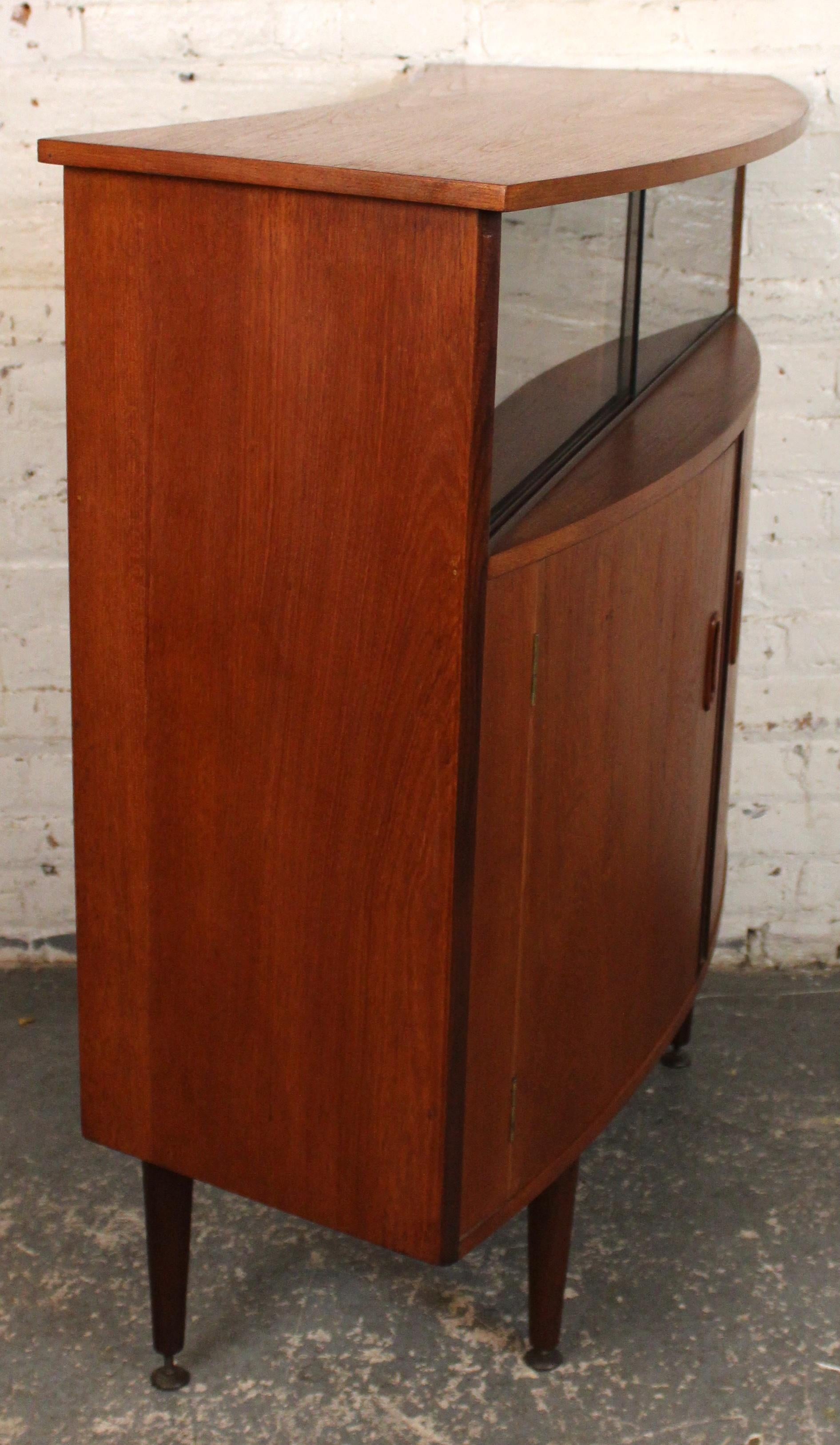 Ein auffälliger Mid-Century Modern-Cocktailschrank aus reichlich getöntem Teakholz mit einer elegant geschwungenen Front, die die natürliche Maserung des Holzes zur Geltung bringt. Das Design lässt sich öffnen und gibt den Blick frei auf durchdacht
