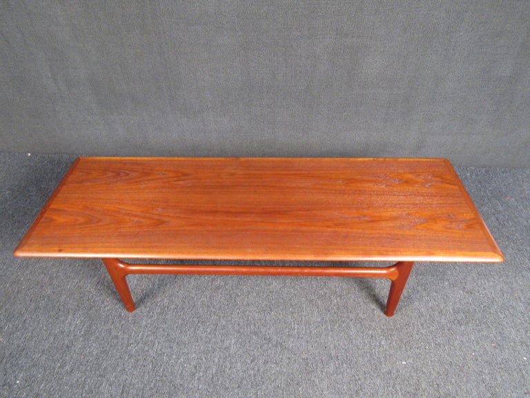 Elegant Danish vintage modern teak coffee table with beautiful wood grain throughout. This vintage rectangular coffee table boasts cylindrical legs connected by stretchers ensuring maximum sturdiness. The perfect addition to any home, business, or