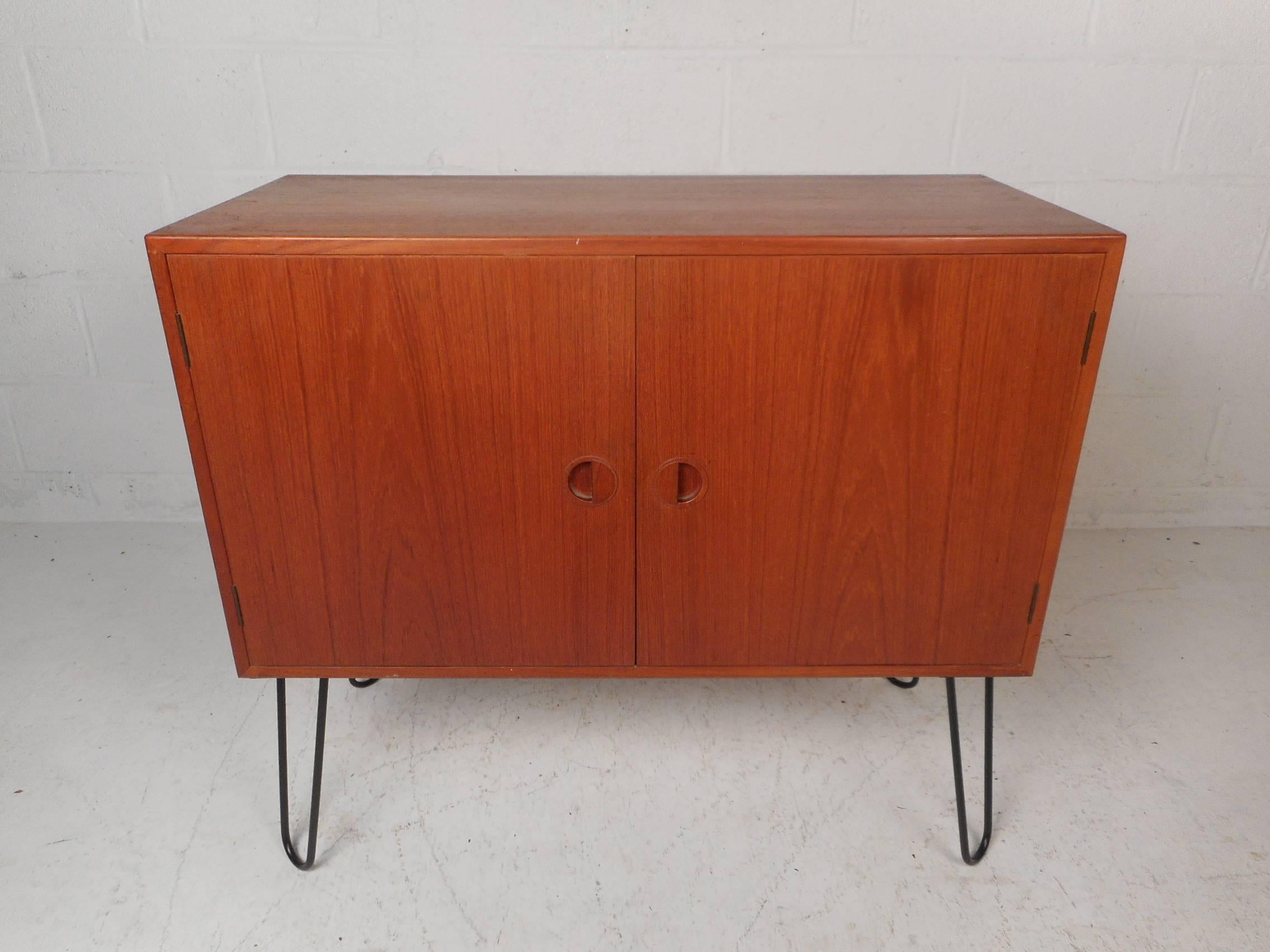 This stunning vintage modern case piece features two cabinet doors with half moon recessed pulls hiding two large compartments with shelves. This Danish modern cabinet sits on top of sturdy black metal hairpin legs. An elegant teak finish