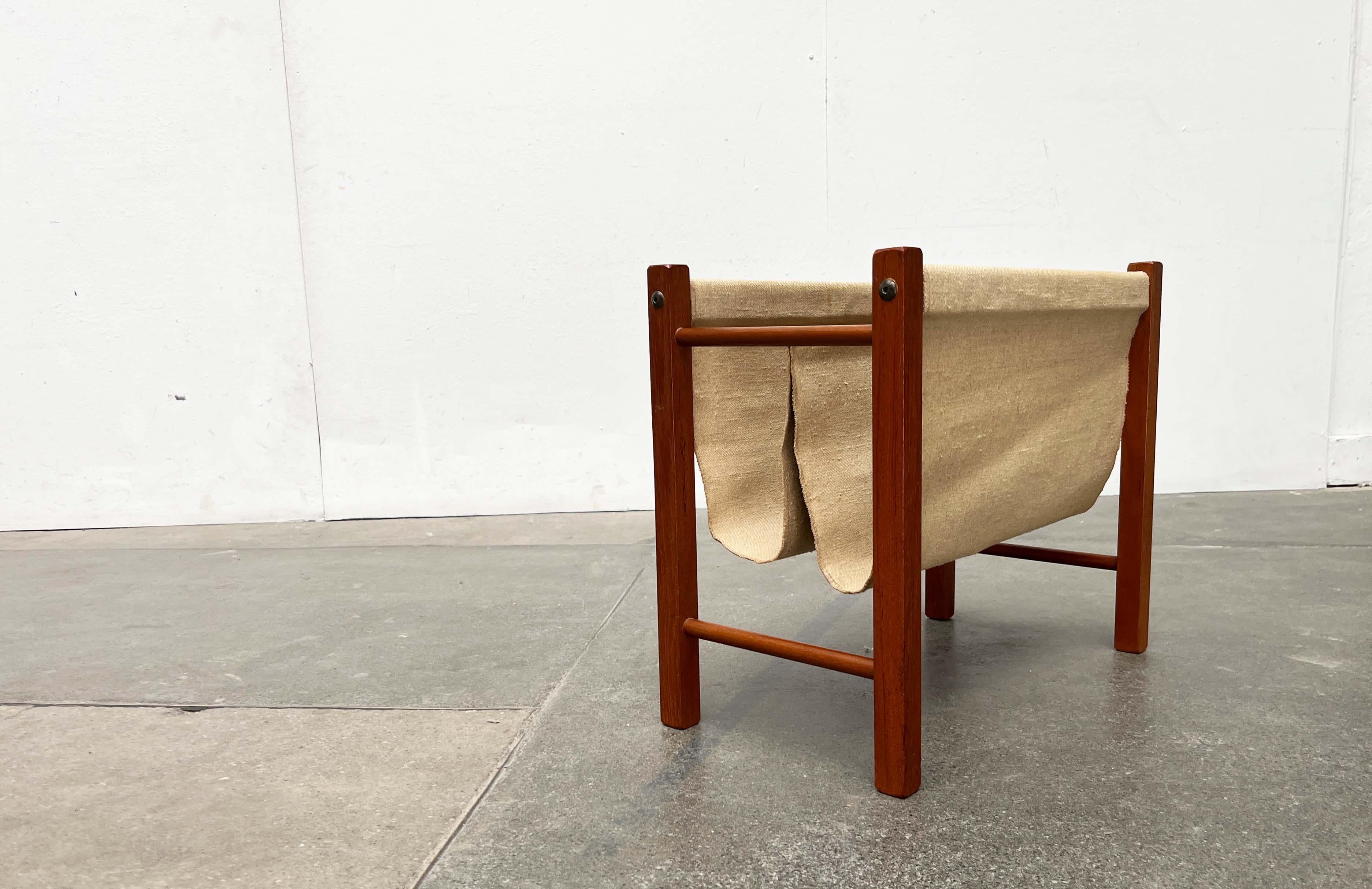 Elegant mid-century teak magazine rack.

The finely crafted frame made of solid teak wood elements is striking. The actual shelf consists of a coarse canvas fabric clamped into the frame. The central bar is made of brass.
