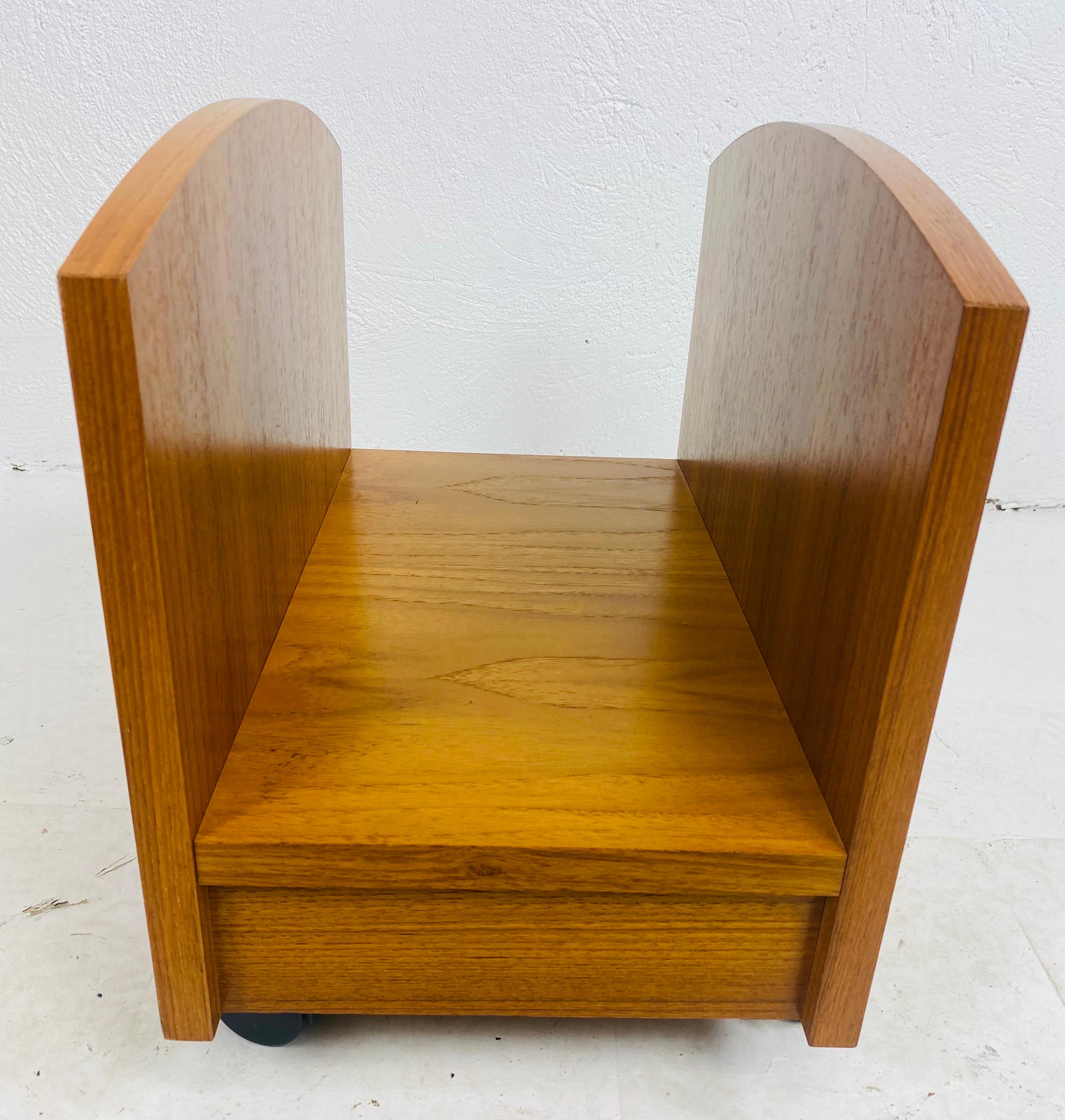 This is a mid century modern danish magazine rack. This teak magazine rack rests on for black wheels. This teak magazine rack has a soft golden tone to the finish. This piece was made in Denmark circa 1960.