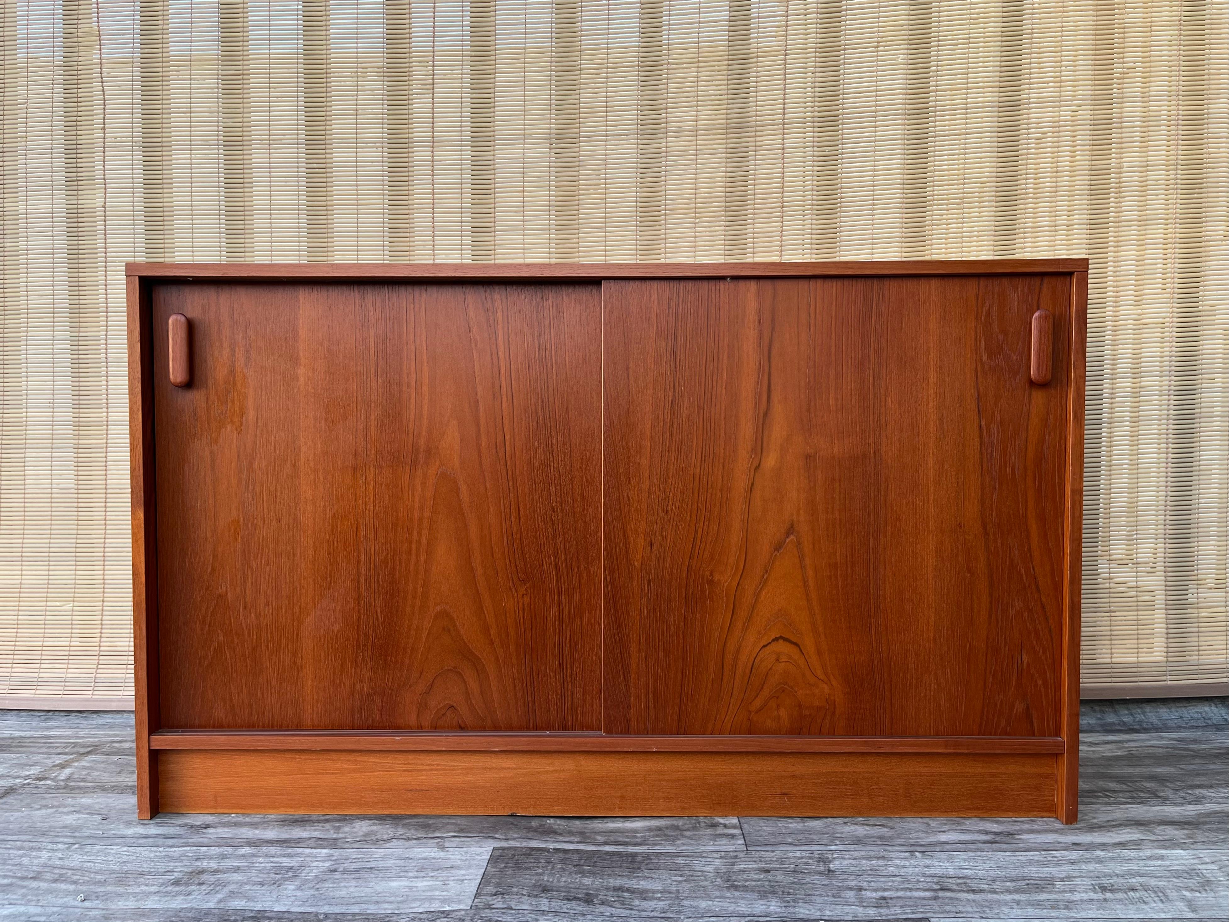 Vintage Mid Century Modern Danish Teak Sideboard/ Credenza. Circa 1970s
Features a sleek minimalist Mid Century Modern Scandinavian Design, a wonderful teak wood grain, two sliding doors, adjustable shelves, and a compact size to fit in any small