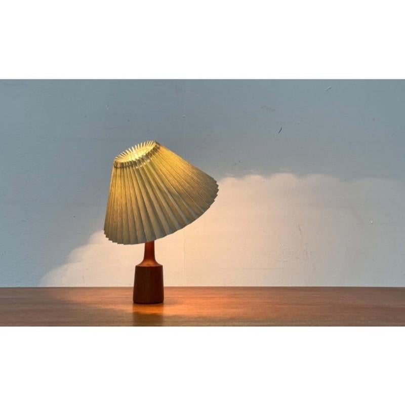 Danish table lamp with a teak base from the 1960s.

The conical pleated shade sits directly on the bulb with a clamp. This clamp allows for manual adjustment of the shade. The base is a simple rotating body made of solid teak.
The lamp is in very