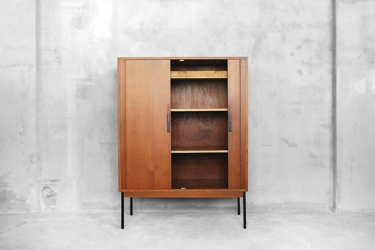 This cabinet was manufactured in Denmark during the 1950s. It is made from wood and teak veneer and has two hidden tambour doors with modern handles. The legs are made from metal. The cabinet has two adjustable shelves and one drawer inside.

This