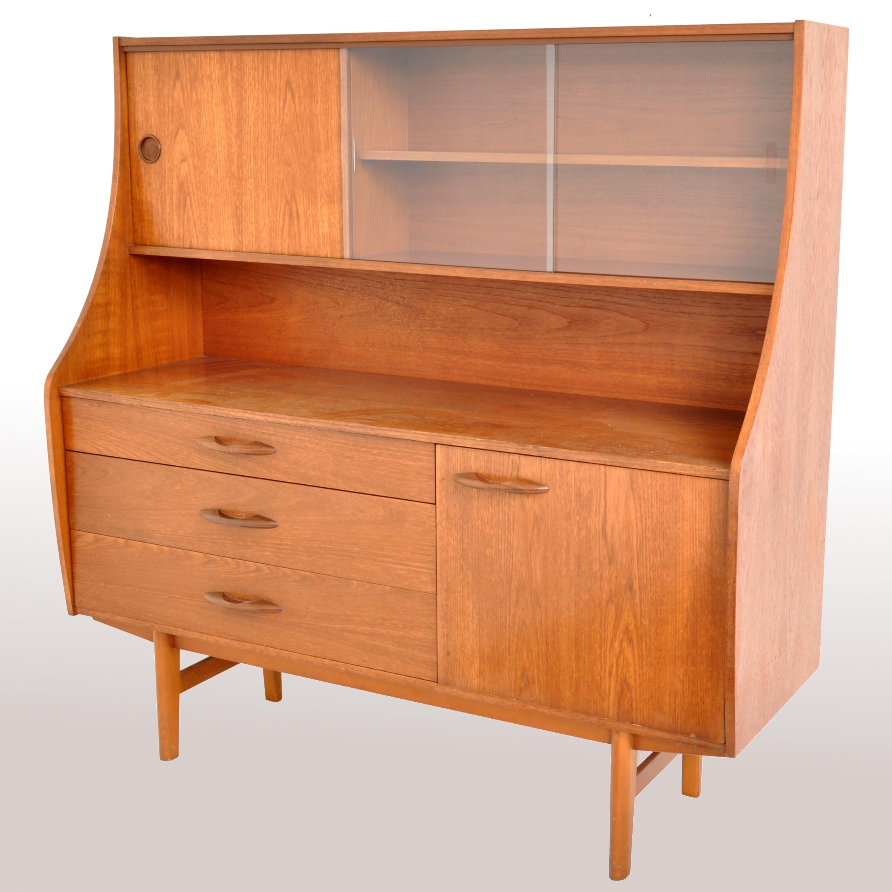 An original 1960s teak credenza having a sliding cabinet door to the left and twin sliding glass doors enclosing a single shelf to the right. Below is a recessed cavity above a bank of three graduated drawers to the left and a hinged cabinet door