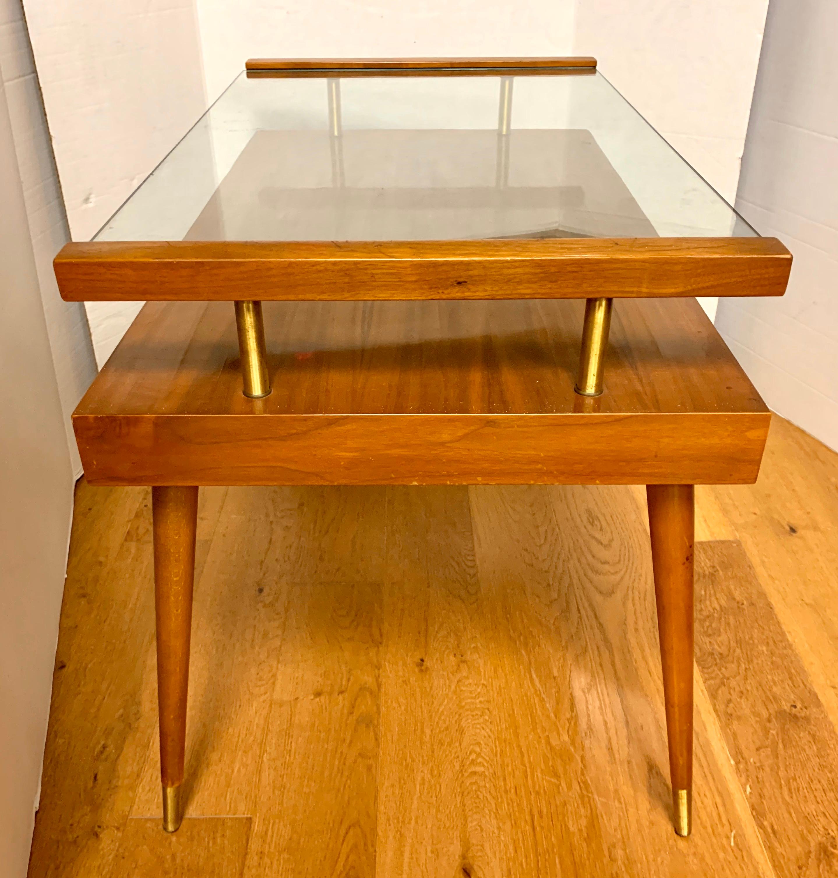 Resting on atomic style legs, this midcentury two-tiered accent table a glass top that slides out and the aforementioned two levels of space. Legs also feature brass caps. An iconic, true period table for the discerning collector of fine midcentury