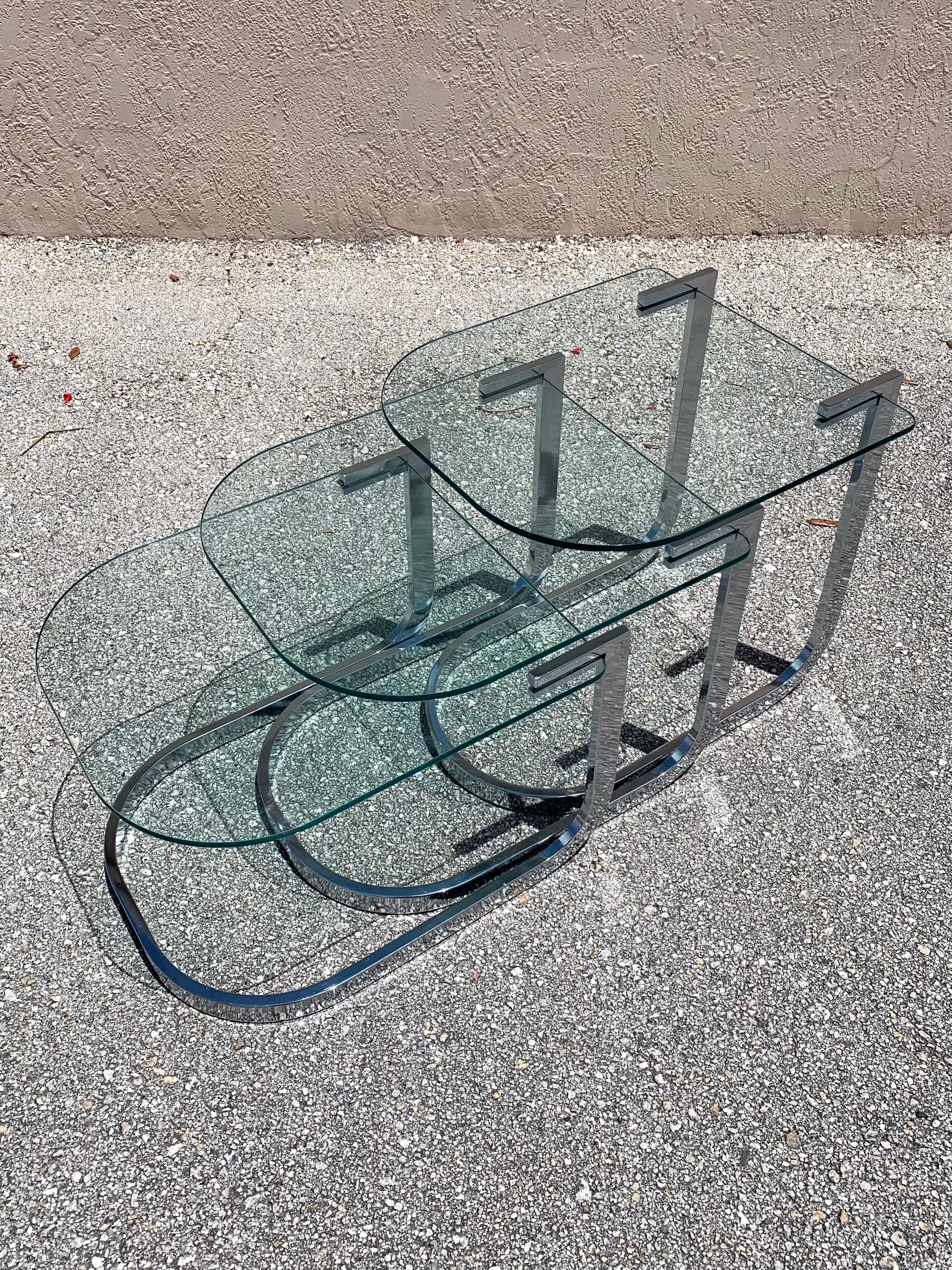 A set of 3 chrome and glass nesting tables by Design Institute of America. Clean and flowing design that looks good stacked together or spread out. Would make a great multi tiered coffee table as well. Flowing chrome frame with a floating glass