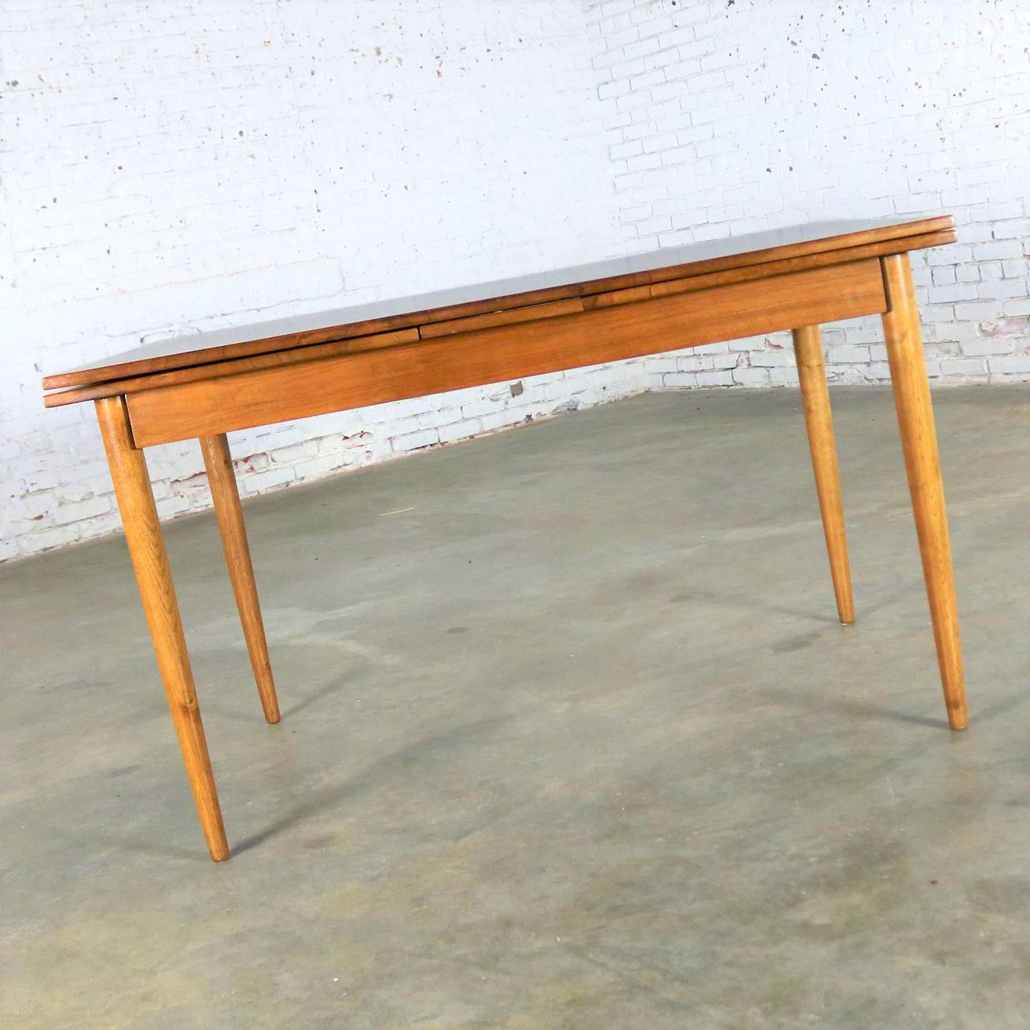 Handsome Mid-Century Modern draw leaf and extending dining table after Conant ball. It is in good vintage condition. We believe it to have been refinished at some point in its life and there is a small spot of missing veneer on the bottom edge on