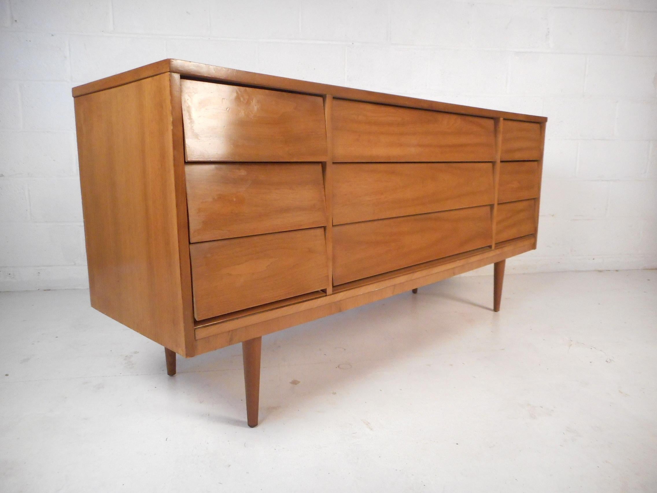 This stylish set of Mid-Century bedroom furniture features a nine-drawer dresser and two nightstands with matching walnut wood grain throughout. Sleek angular drawer faces with dovetail construction ensure ample storage space. Sturdy construction