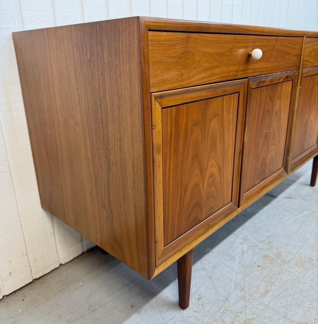 Questa inserzione si riferisce a una credenza in legno di noce della Dichiarazione di Drexel, un'opera moderna di metà secolo. Caratterizzato da un design lineare, due cassetti nella parte superiore con pomelli originali in vetro al latte, quattro