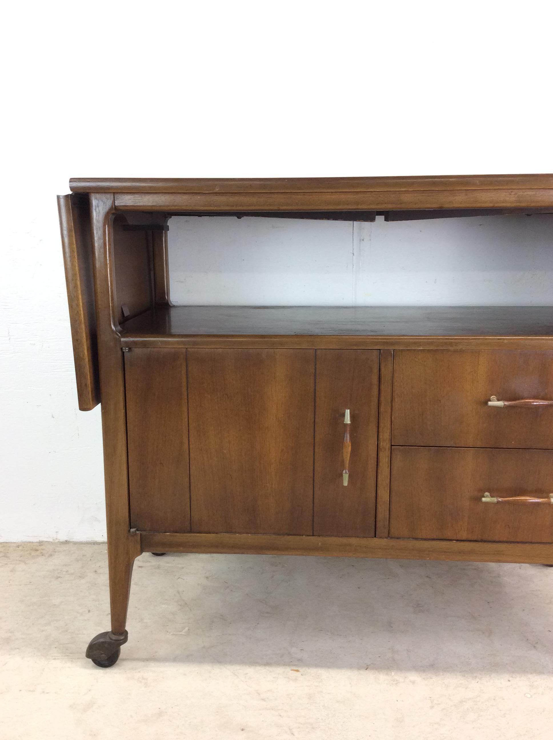 This mid century modern bar cart / wheeled server features hardwood construction, original walnut finish, storage cabinet, two dovetailed drawers with brass accented hardware, black lacquer top, two drop leaf extensions, and tall tapered legs with