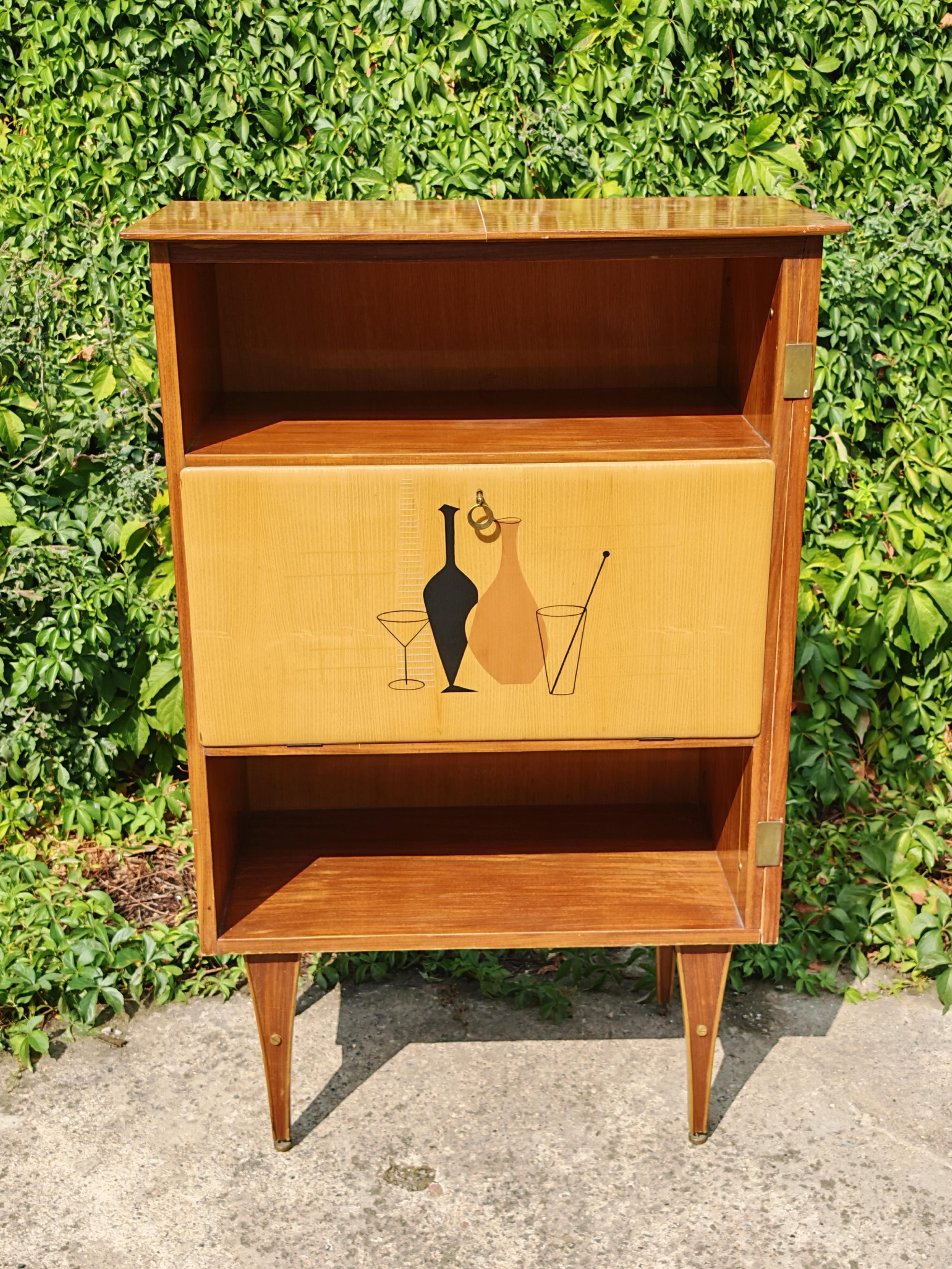 In this listing you will find a striking Mid Century Modern Bar Cabinet. It is done in mahogany veneer, with brass accent and glass shelf hidden inside the cabinet. The cabinet door feature a beautiful inlay depicting bottles and glasses. It also