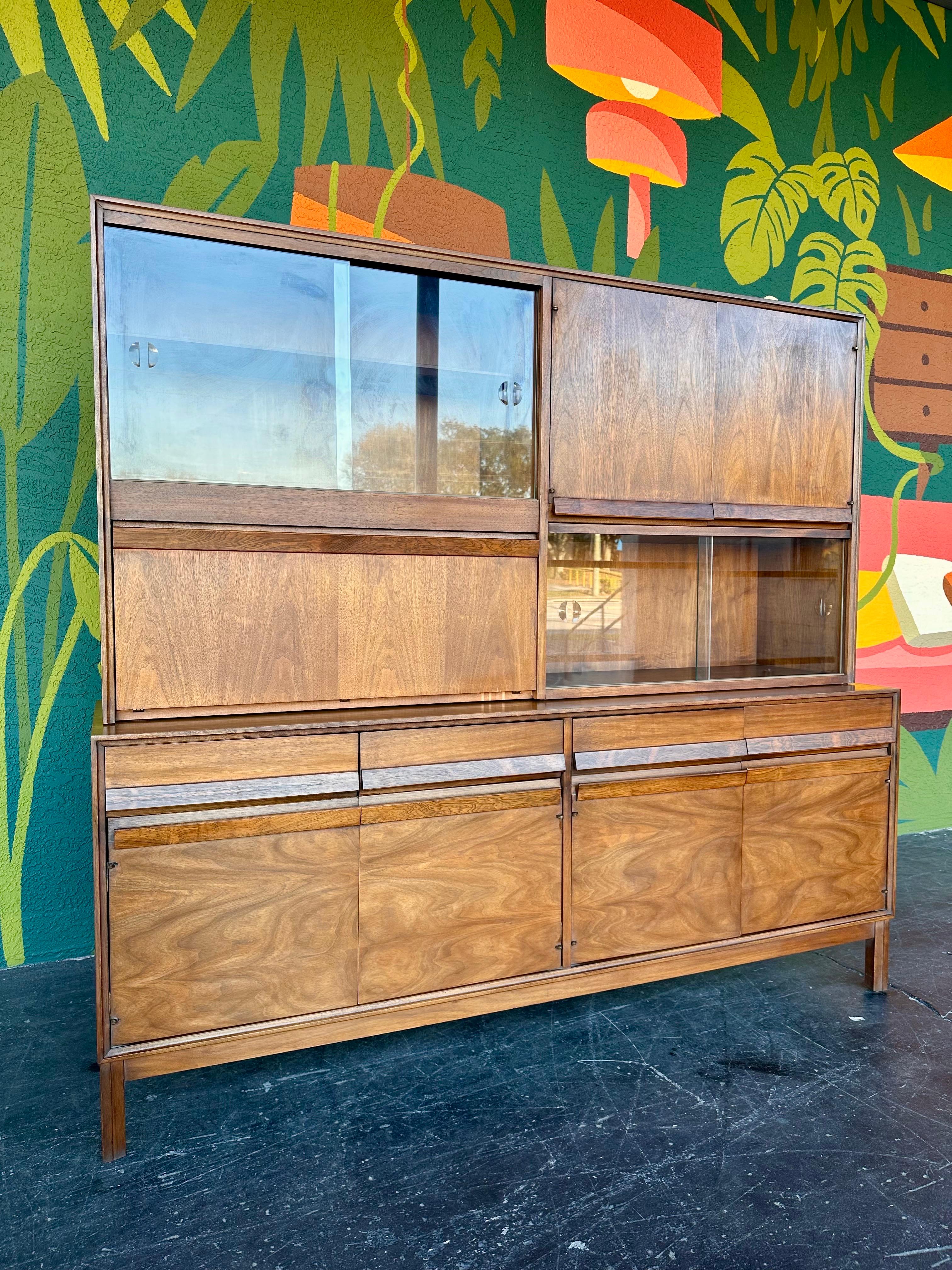 Mid Century Modern Walnut and Glass two piece dry bar china hutch by American of Martinsville. Upper section features two sets of sliding glass doors, a drop down bar cabinet, and closed storage. Lower credenza section has four drawers and two