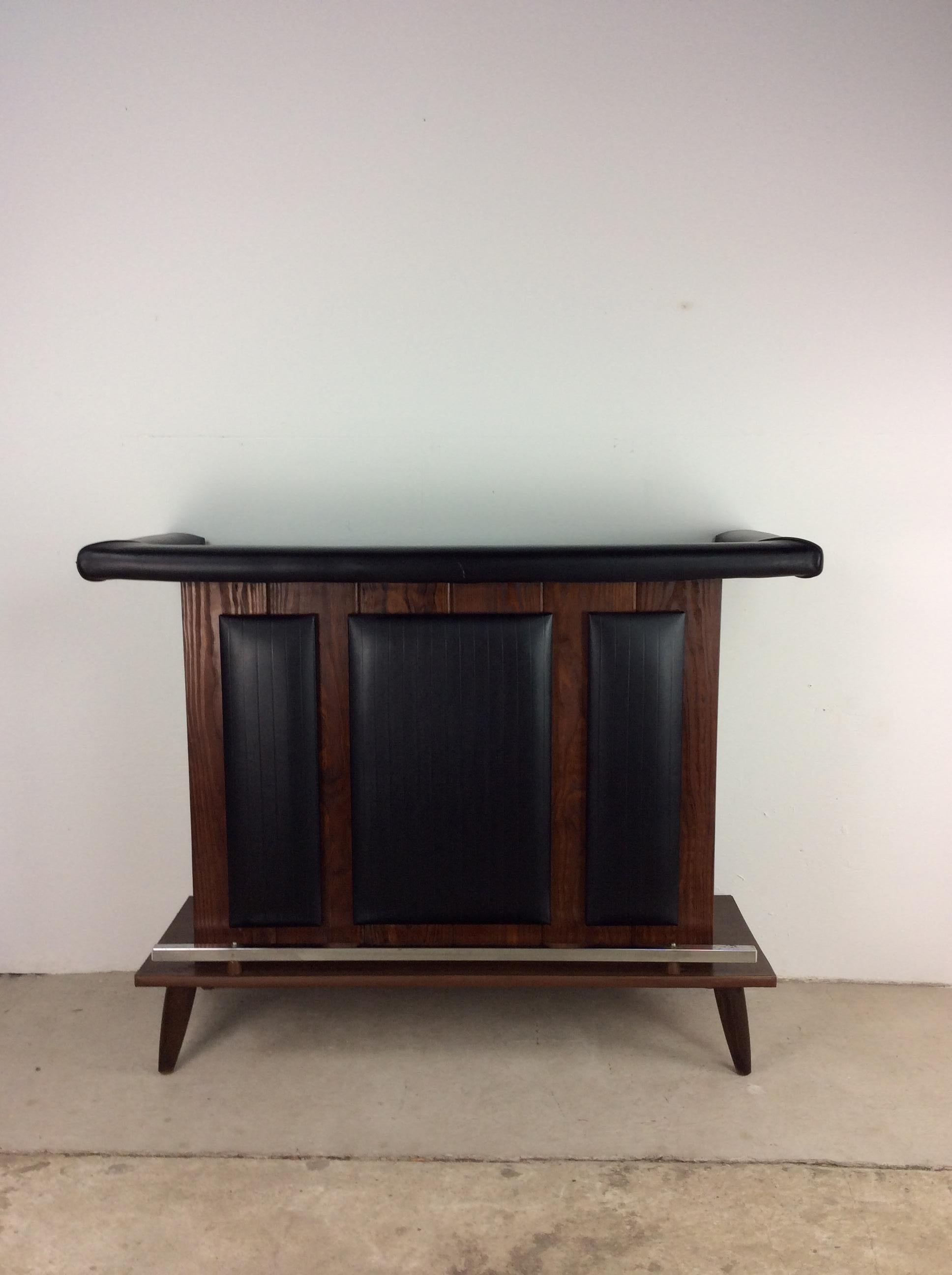 This Mid-Century Modern dry bar features hardwood construction, original walnut finish, black vinyl counter edge with black vinyl accents on the bar front, tapered legs, open shelving in the back and a chrome kick rail.

Dimensions: 51.5w 19d