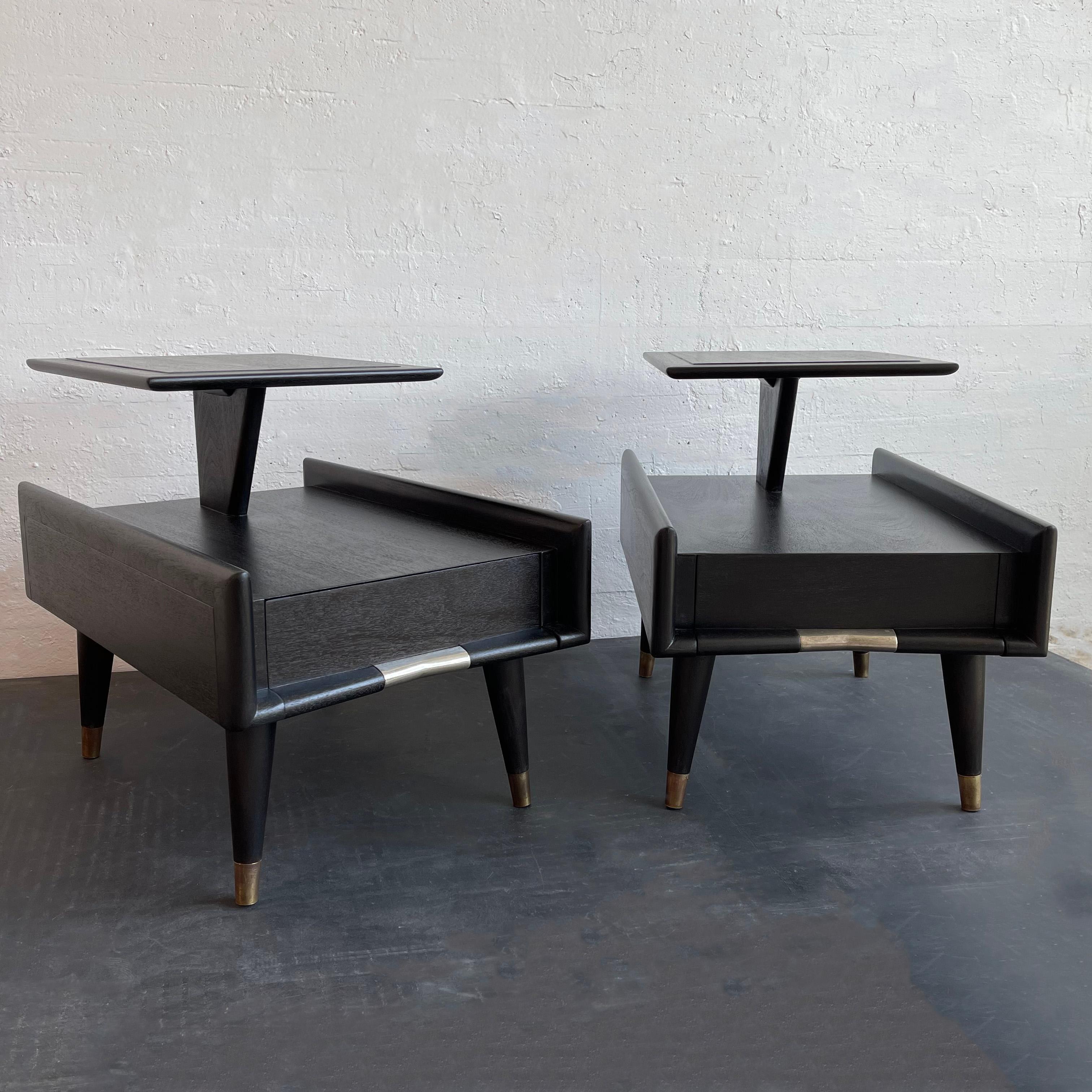 Striking pair of atomic, mid-century modern, ebonized mahogany, stepped end tables or nightstands produced by Gordon’s Inc. Furniture company. The tiered tables feature tapered legs with brass sabots and aluminum drawer accents. The lower tier