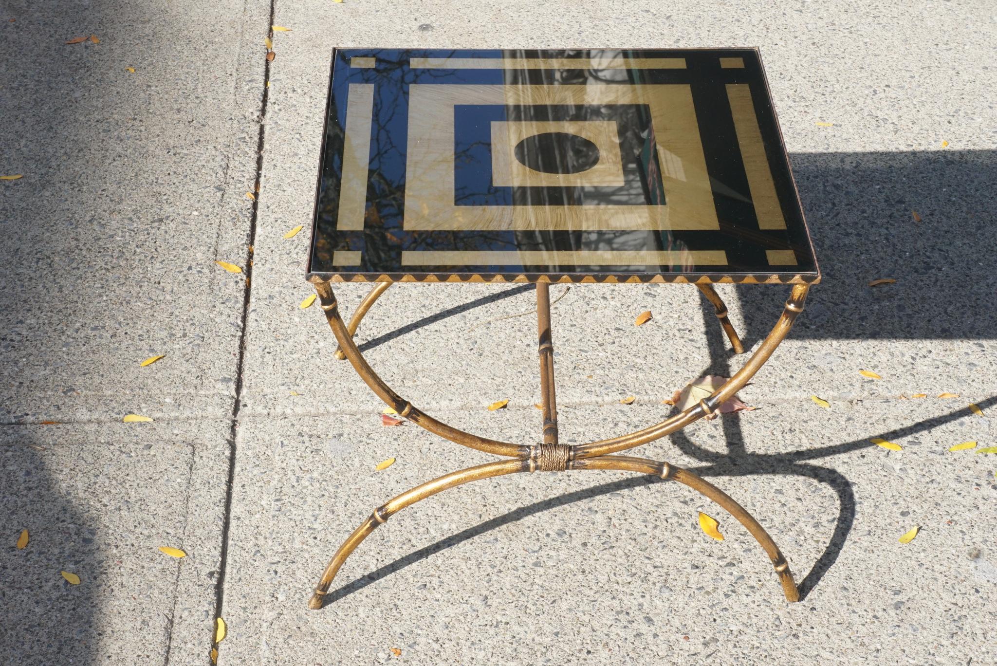 Made in America circa 1955 this low wrought iron table is made to resemble bamboo and is then gilded and painted with a wavy decorative border along the apron. Set within the frame of the apron is a reverse glass painted and gilded top. The design