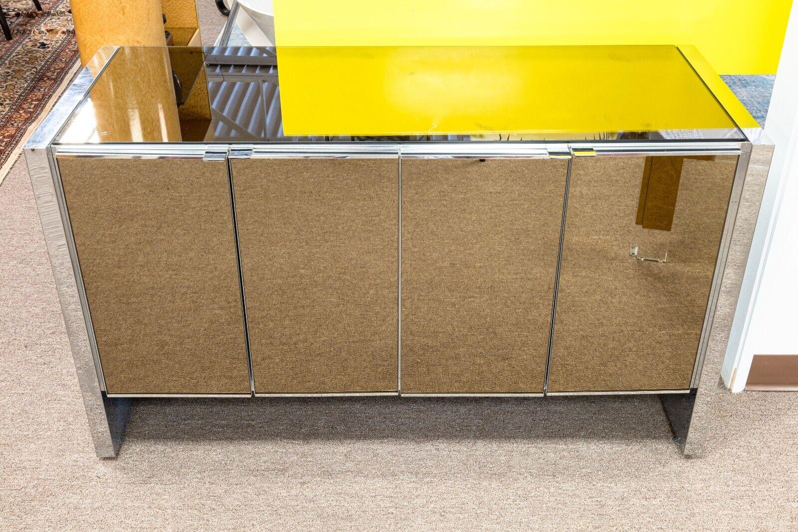 An Ello mirrored chrome credenza. This lovely credenza features a very lovely mirrored design, with mirrors wrapping around the front, sides, and top of this piece. Aside from this, there is also chrome detailing throughout the piece. This credenza