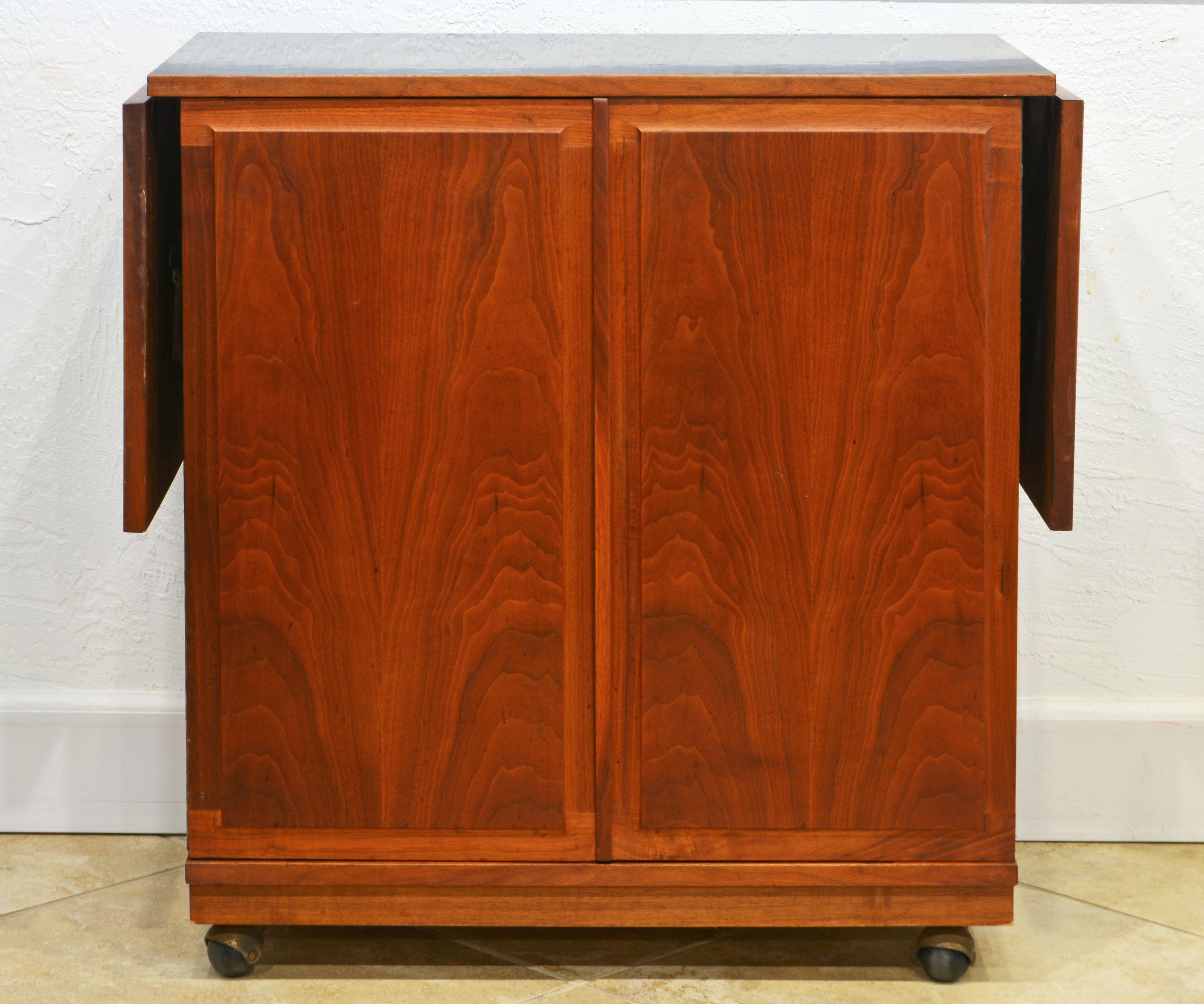 This Mid-Century Modern walnut bar cart and cabinet features a wood edged black laminate top and drop leaves, two paneled figured walnut doors on piano hinges and with an innovative opening grip all in wood resting on wheels partly concealed by a