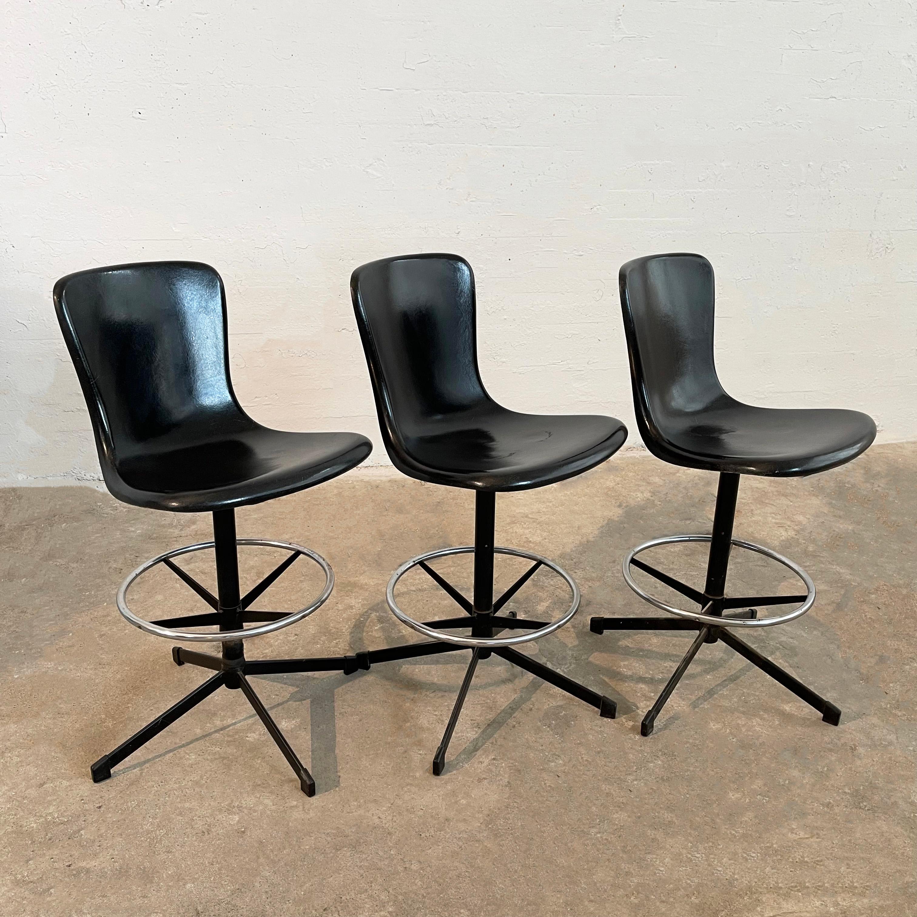 Set of three, mid-century modern, counter height, swivel stools by Cosco feature black fiberglass shell seats with backs on black metal pedestal bases with chrome ring footrests. The stool's footprint is 22 inches diameter and the footrest is 15