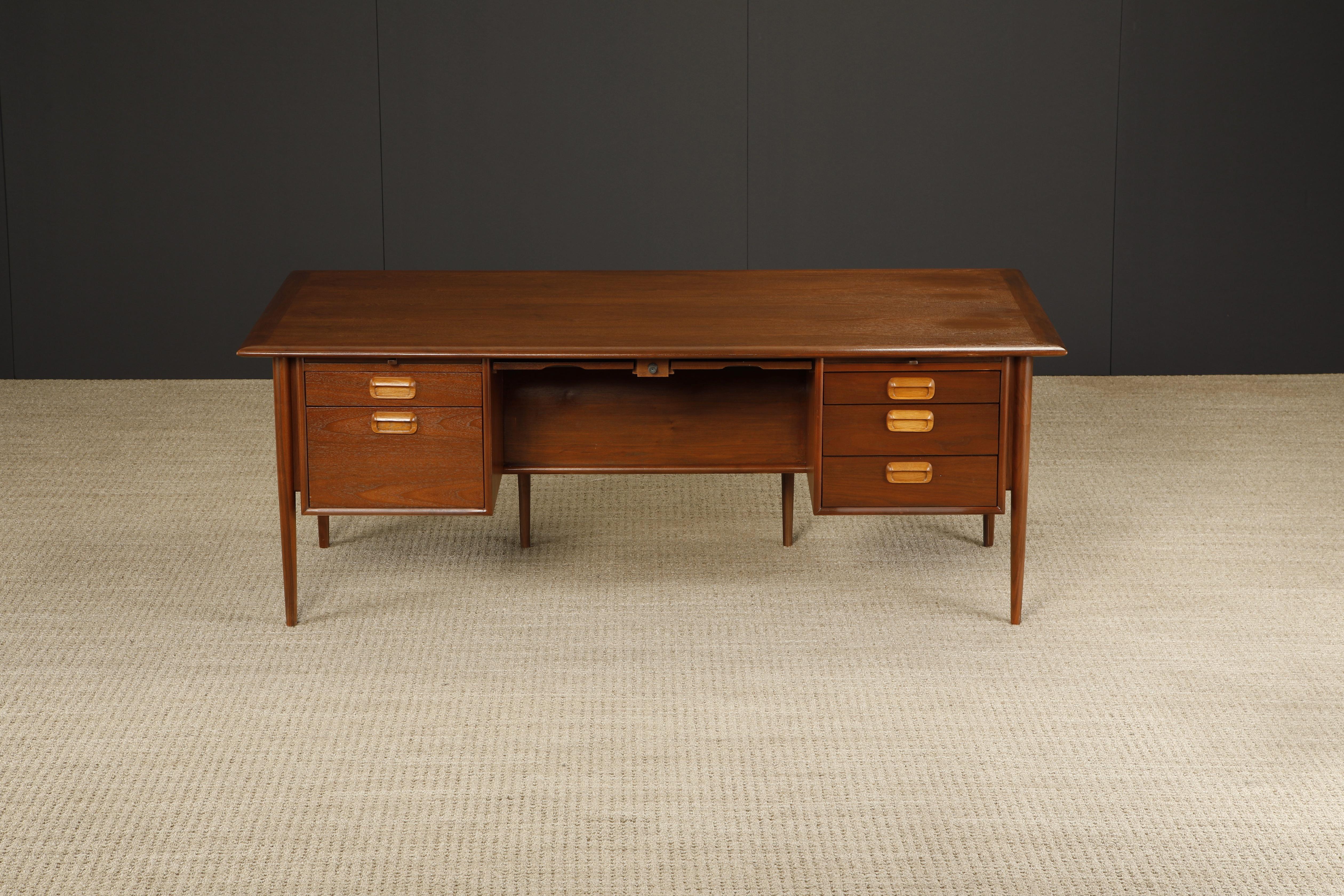This Mid-Century Modern executive desk, circa 1960s, crafted from walnut features a Arne Vodder styled floating top over six legs, with contrasting oak handles, four pull out trays (two in the center flanked by two on the ends), and five drawers.