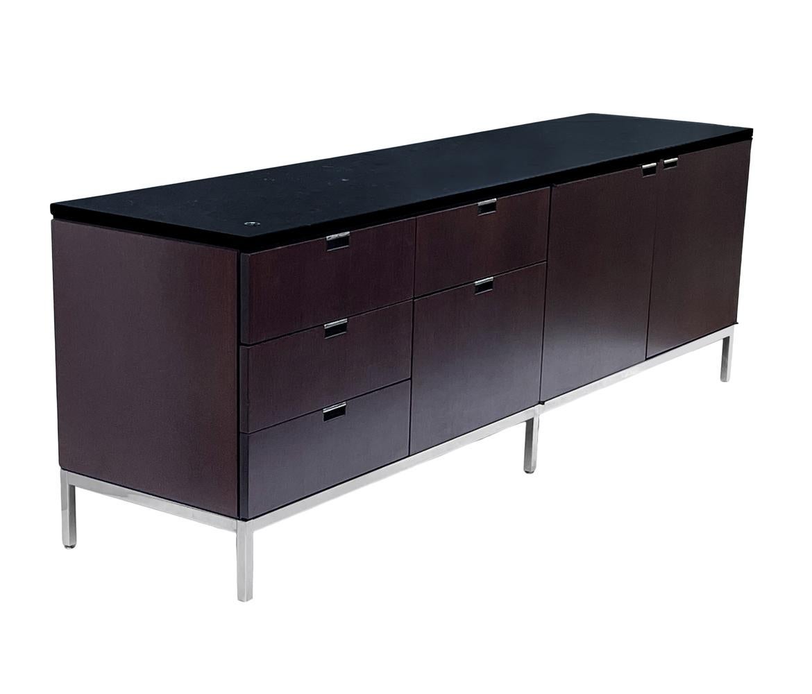 A sleek modern classic cabinet designed by Florence Knoll and produced by Knoll. It features a dark mahogany case, stainless steel legs and a black marble top. We currently have two matching credenzas in stock and they're priced each.