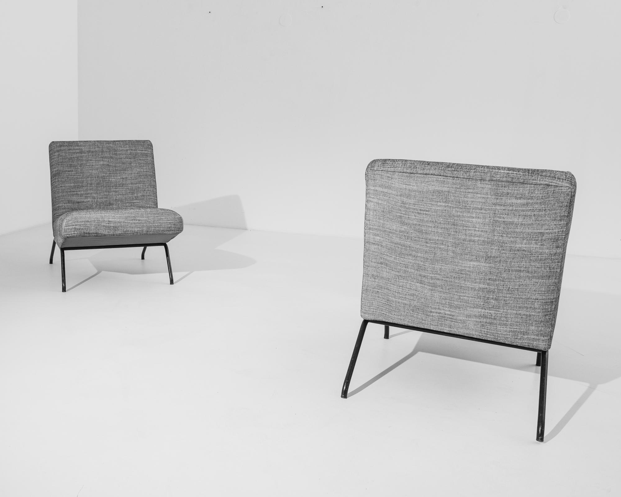 A pair of French mid-century armchairs circa 1960. Upholstered in elegant grey, these modernist chairs feature comfortable, slightly angular seats held up by slender metallic legs of black color. The absence of armrests accentuates their quirky