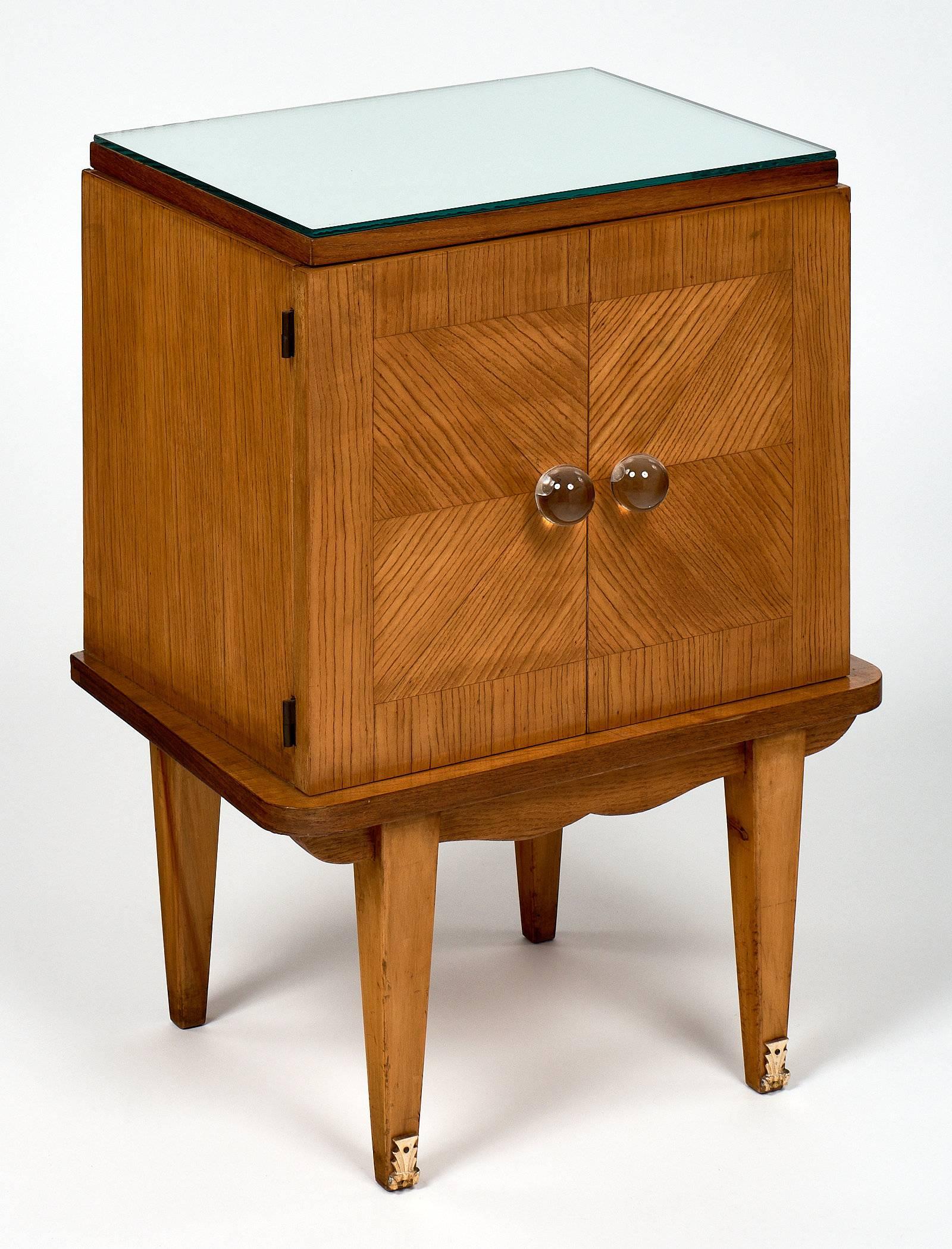 A pair of French Mid-Century Modern side tables, each featuring two doors with clear Murano glass knobs and a single shelf inside. The tables are made of rosewood with mirrored tops. The legs are tapered with gilt brass elements on the front two.