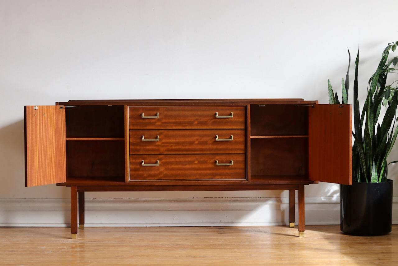 Danish Modern midcentury Tola wood sideboard.
Made by G-Plan in the U.K.
Three dovetailed drawers in the center flanked by cabinets.
Top drawer features dividers.
Each cabinet holds adjustable shelving.
Sleek brass hardware and