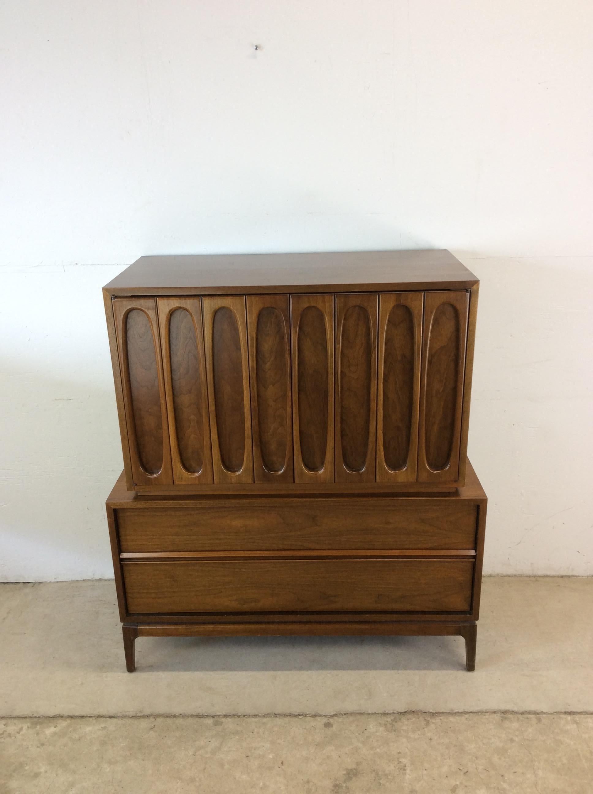 This mid century modern gentleman’s chest features hardwood construction, walnut veneer with original finish, five dovetailed drawers, two cabinet doors with unique sculpted wood detailing, and tall tapered legs.   Matching lowboy dresser and pair